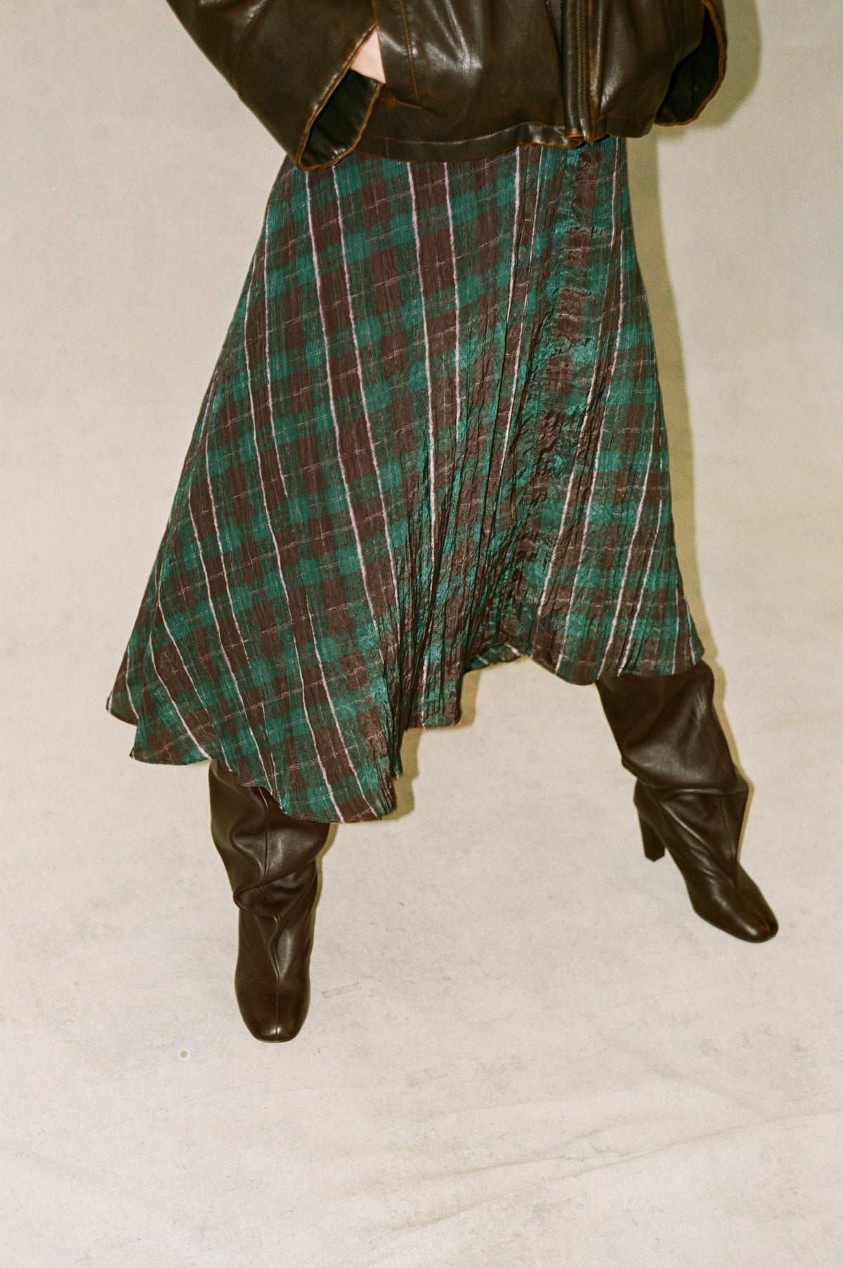 Cropped campaign view of a model in a dark brown leather jacket, flowing teal and burgundy plaid skirt, and tall brown boots.