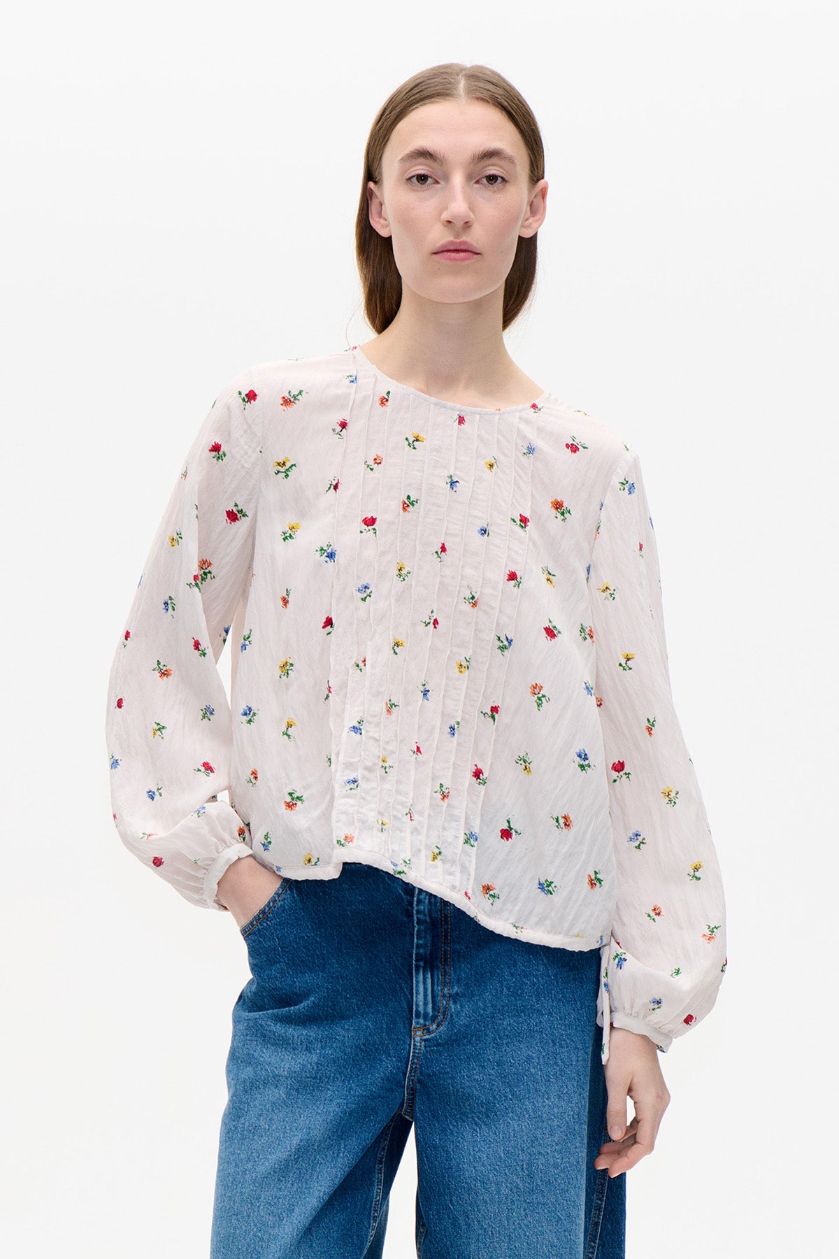 Model wearing a white long-sleeved blouse with small multicoloured floral print and vertical pintuck detailing, paired with blue jeans.