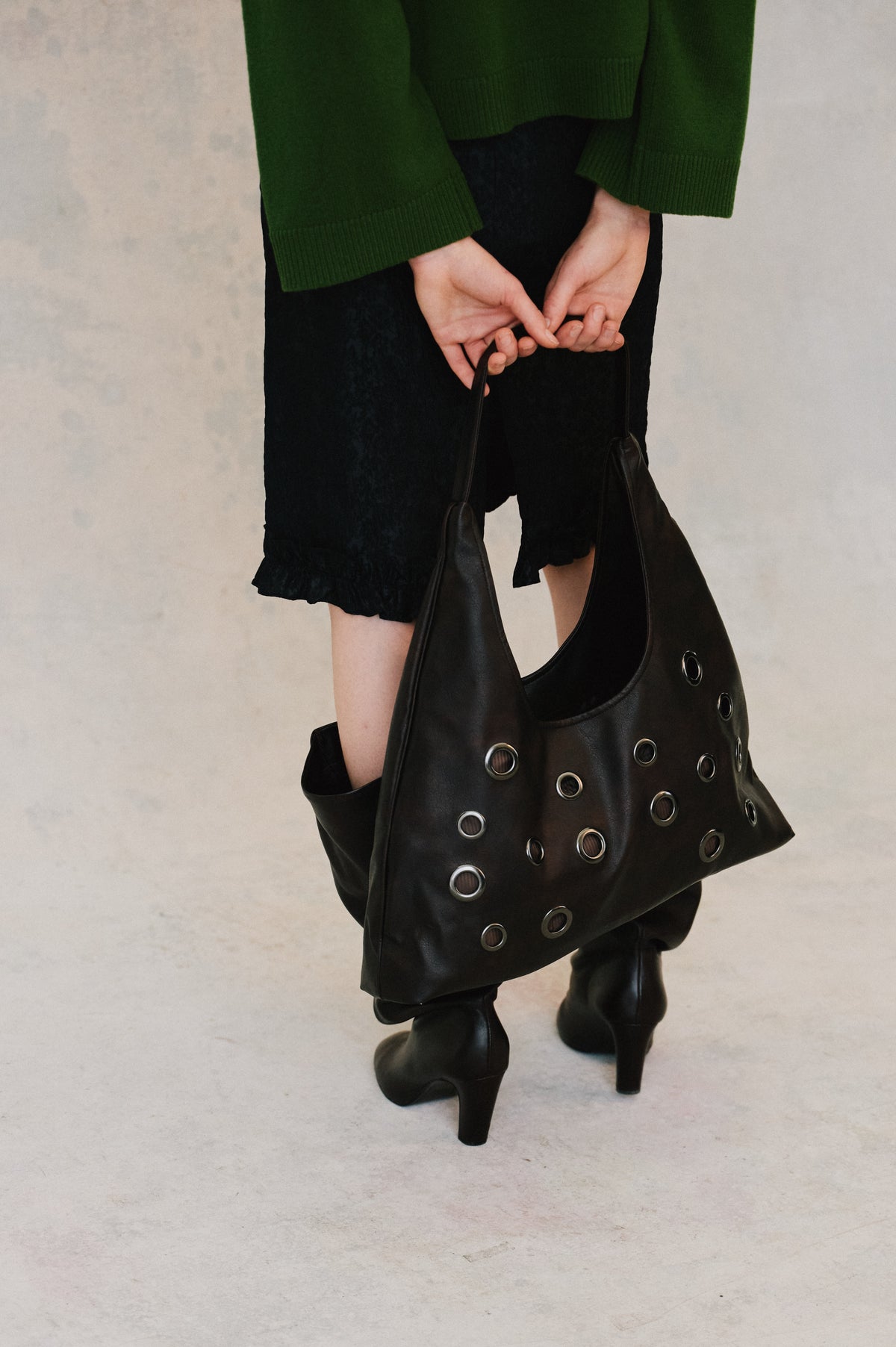 Cropped campaign view of a model in a dark green ribbed sweater and black skirt, holding a large black bag with metal eyelets.