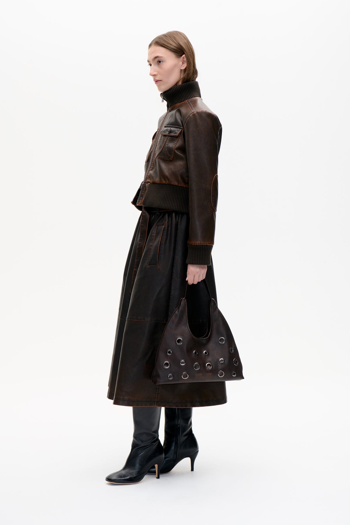 Model in profile wearing a dark brown zip-front jacket with a ribbed black collar, cuffs and hem, a black skirt and boots, carrying a grommet-detailed tote.