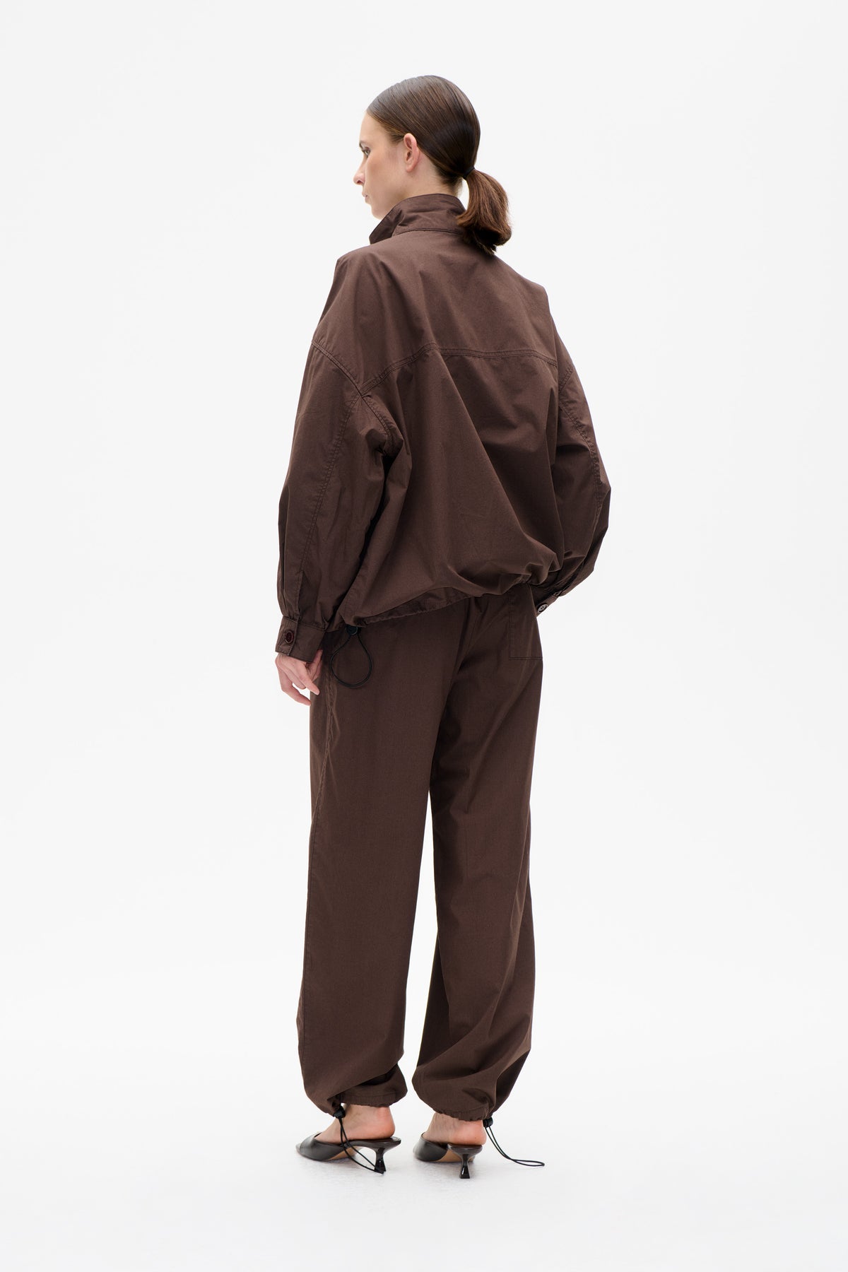Model wearing a dark brown oversized jacket and matching wide-leg trousers, shown from the back with a raised collar and gathered trouser cuffs.