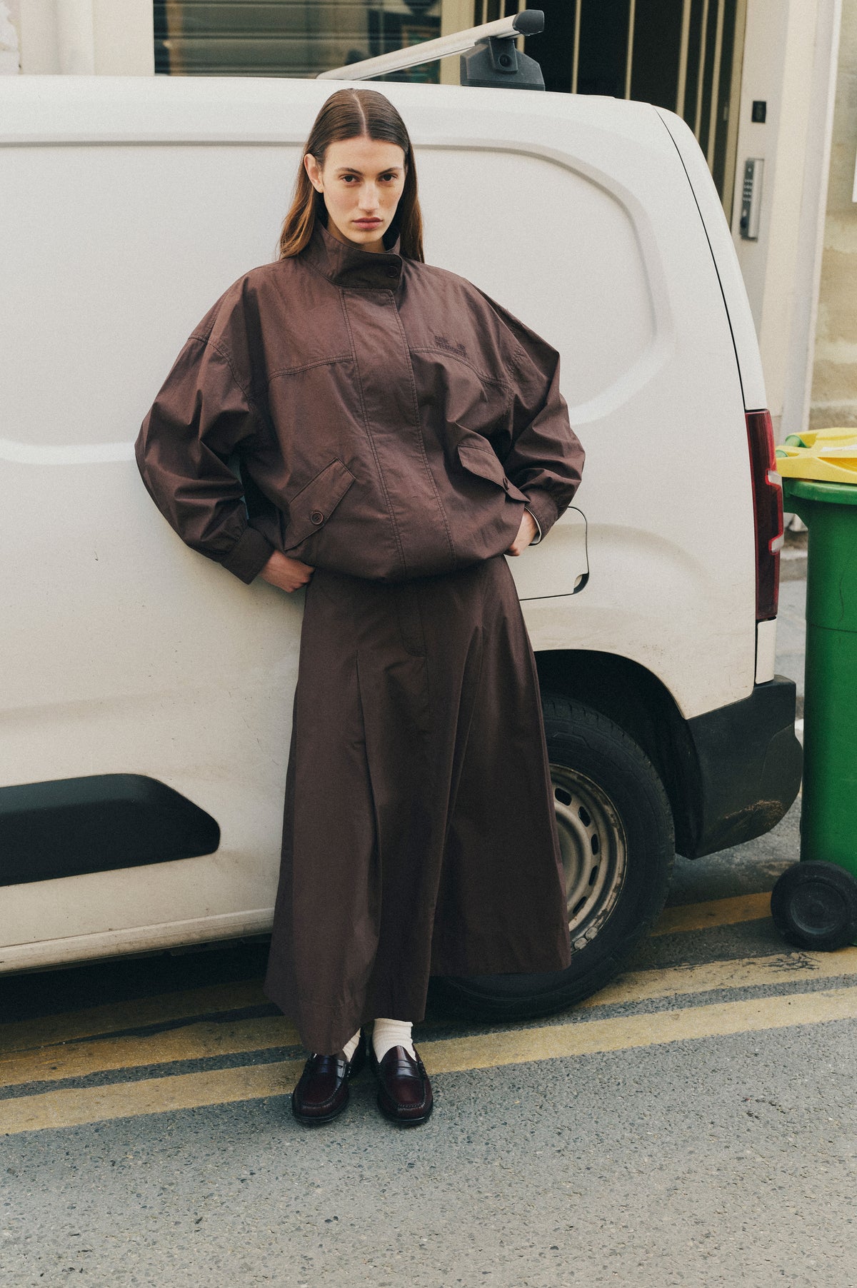 A model poses beside a white van in an oversized dark brown high-collar jacket and matching long skirt, with white socks and loafers.