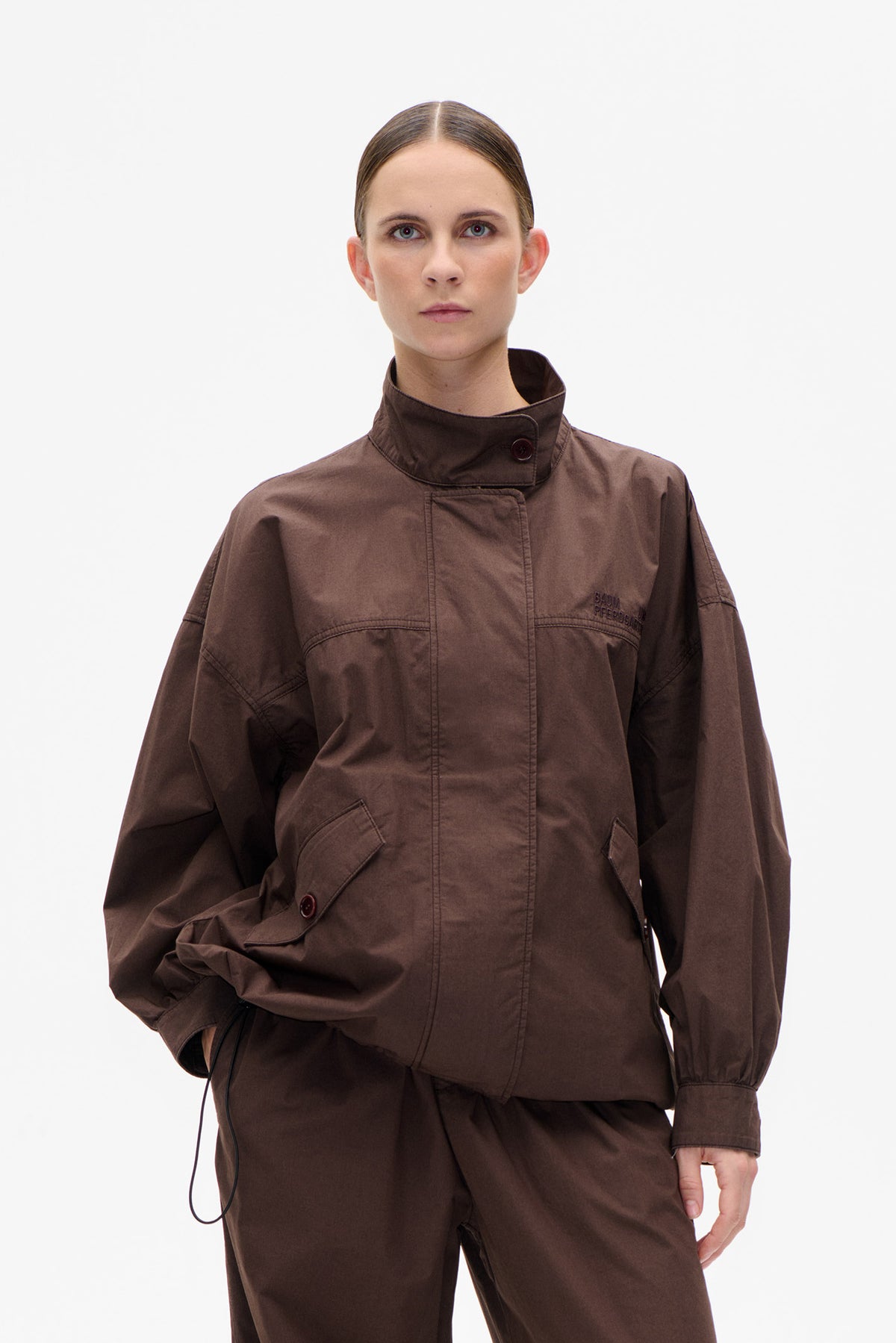 Front view of a dark brown oversized jacket with a high buttoned collar, flap pockets and long sleeves, worn with matching trousers.