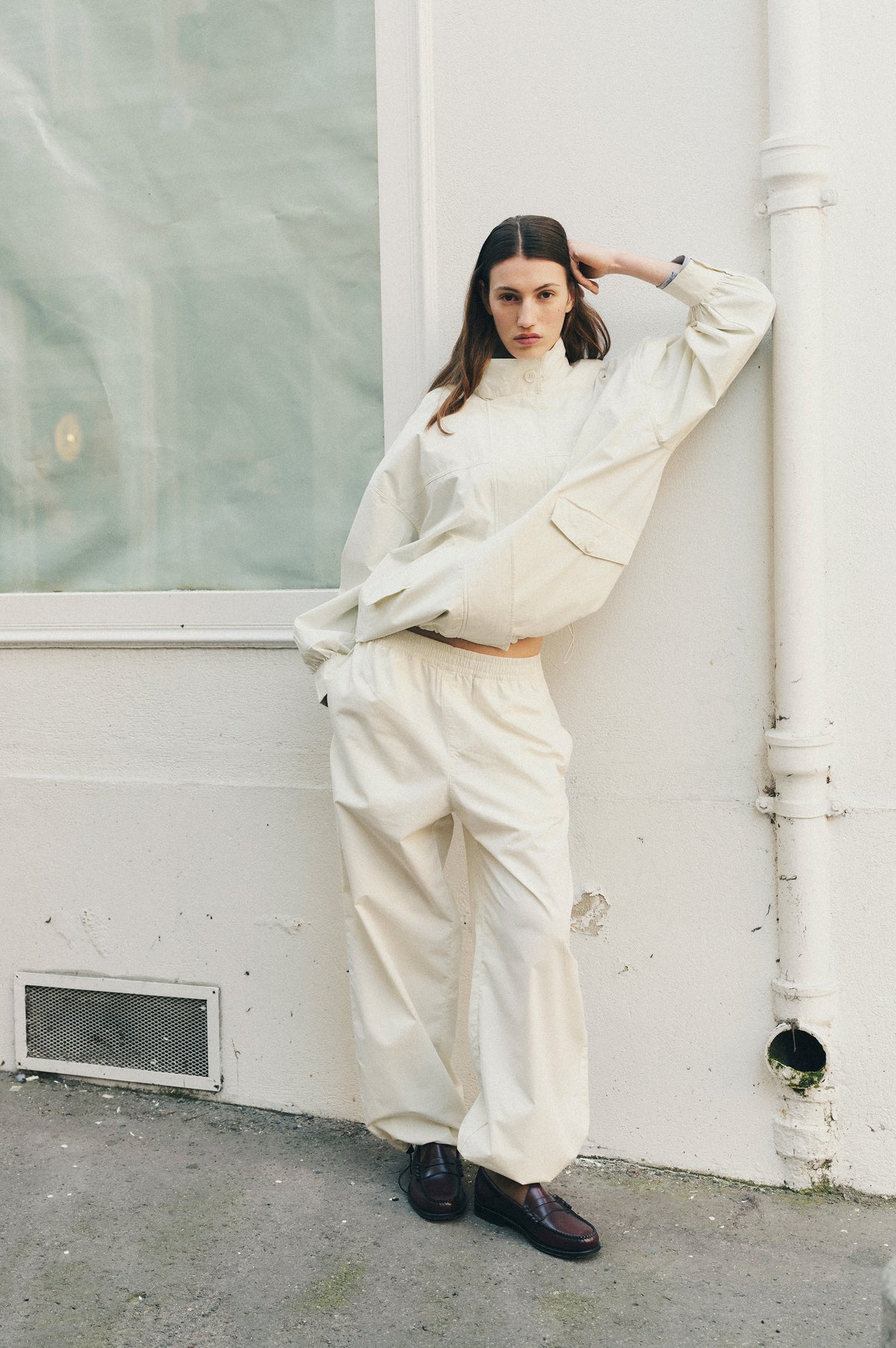 Campaign portrait of a model in an off-white high-neck jacket and loose matching trousers, leaning beside a pale wall.