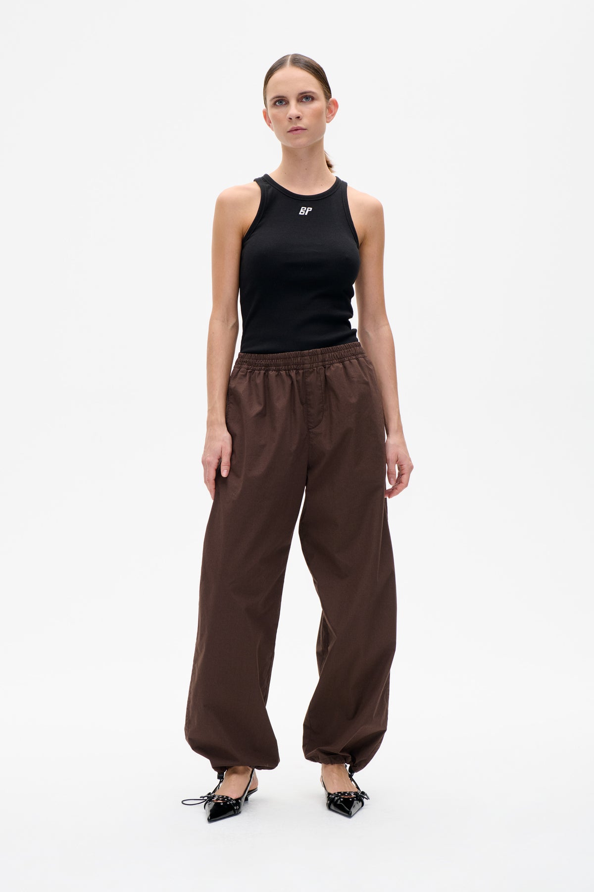 Front view of a black sleeveless tank top with a small white chest logo, paired with dark brown elastic-cuff trousers.