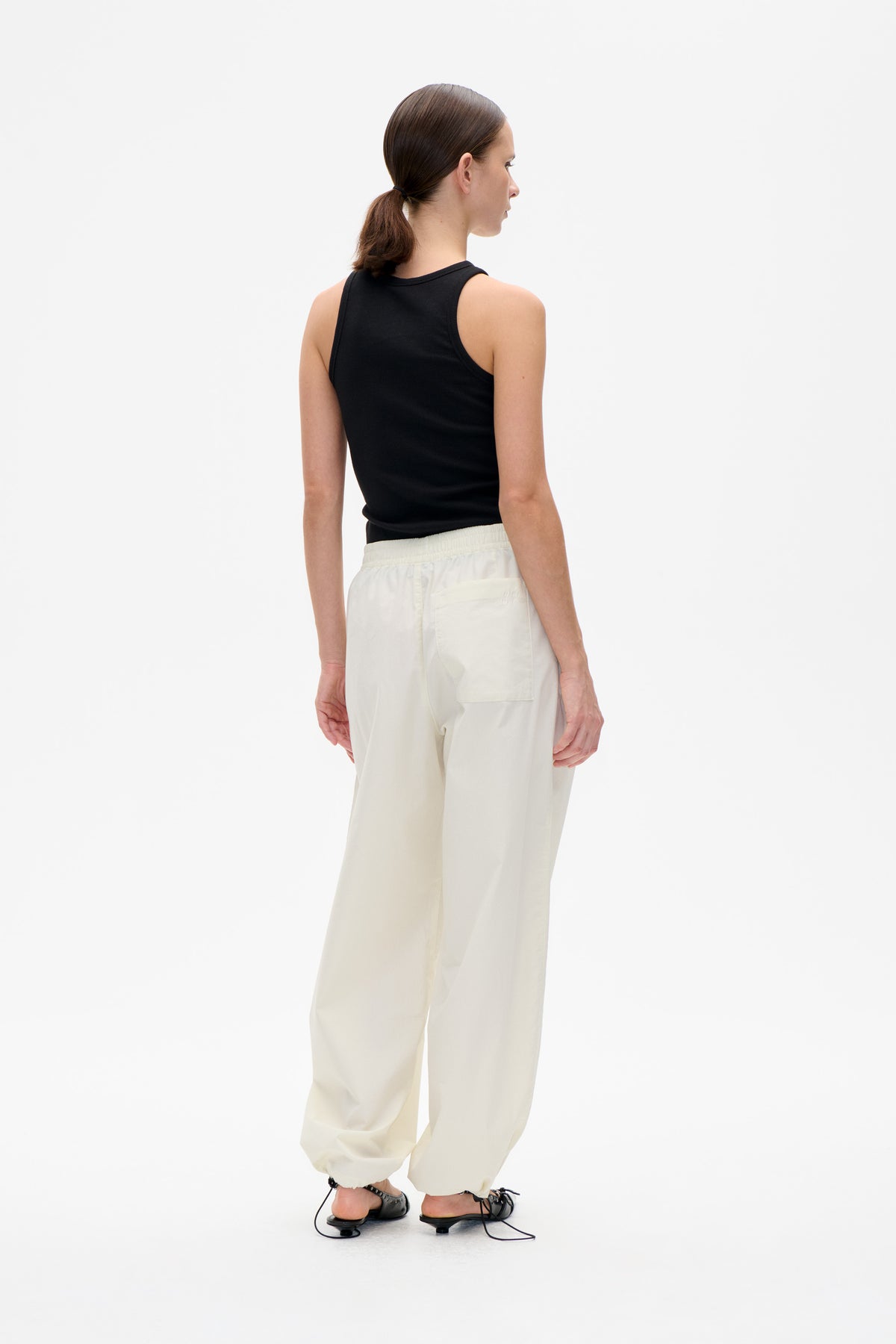 Back view of a model wearing a black sleeveless ribbed top and loose ivory trousers with an elastic waistband and back pocket.