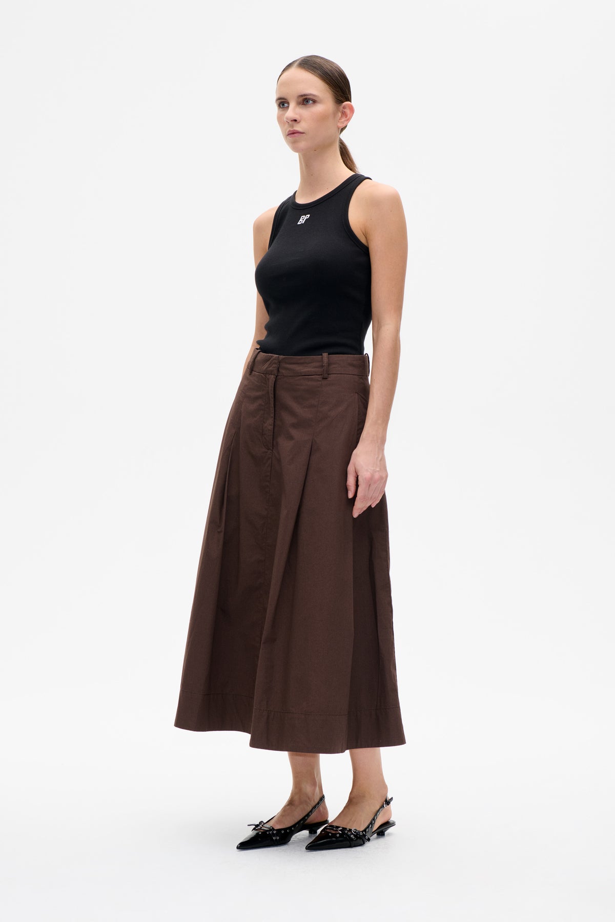 Model wearing a black sleeveless top with a small white BP logo, a brown flared midi skirt and black embellished slingback flats.