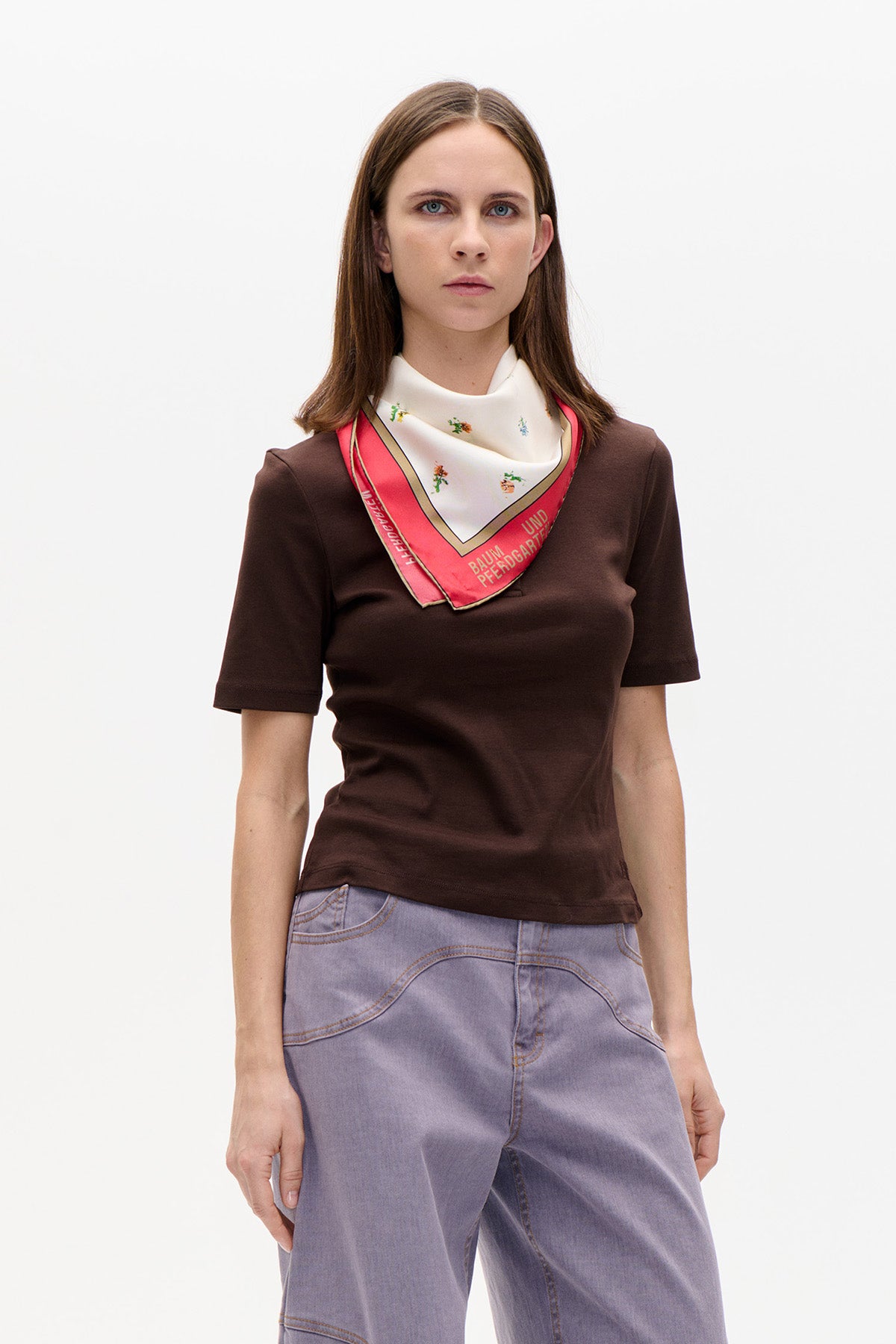 Model wearing a dark brown short-sleeved top, lavender jeans and a cream floral scarf with a coral border and Baum und Pferdgarten lettering.