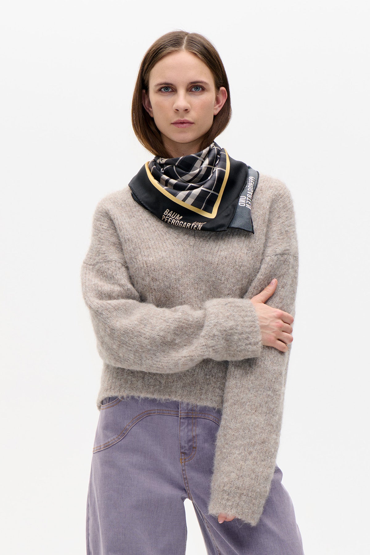 Model wearing a black, cream and grey checked scarf with a gold border over a mottled beige knit sweater and lavender jeans.