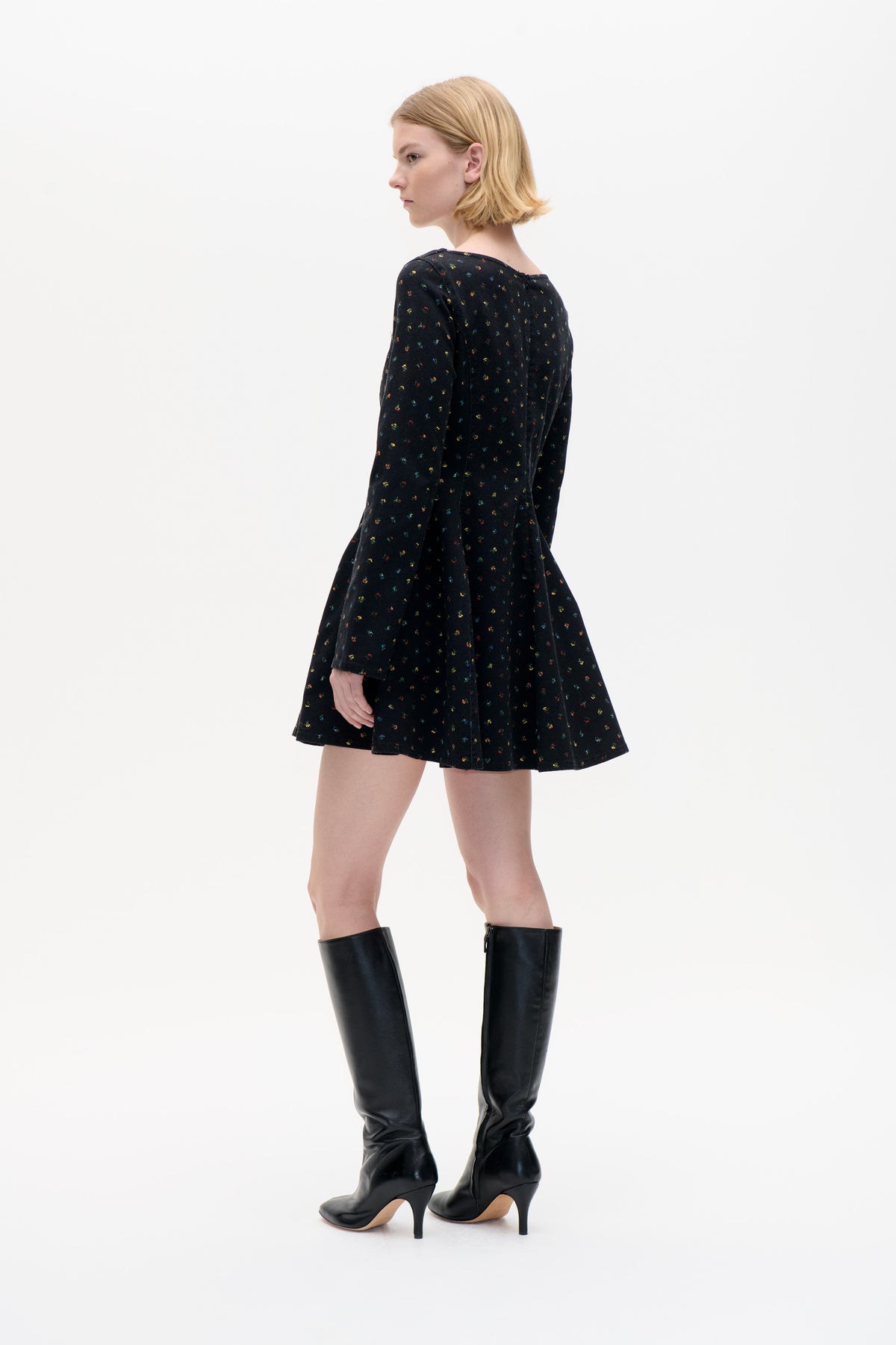 Model wearing a black long-sleeved flared mini dress with tiny multicoloured floral motifs, shown from a back-side angle against a white studio background.