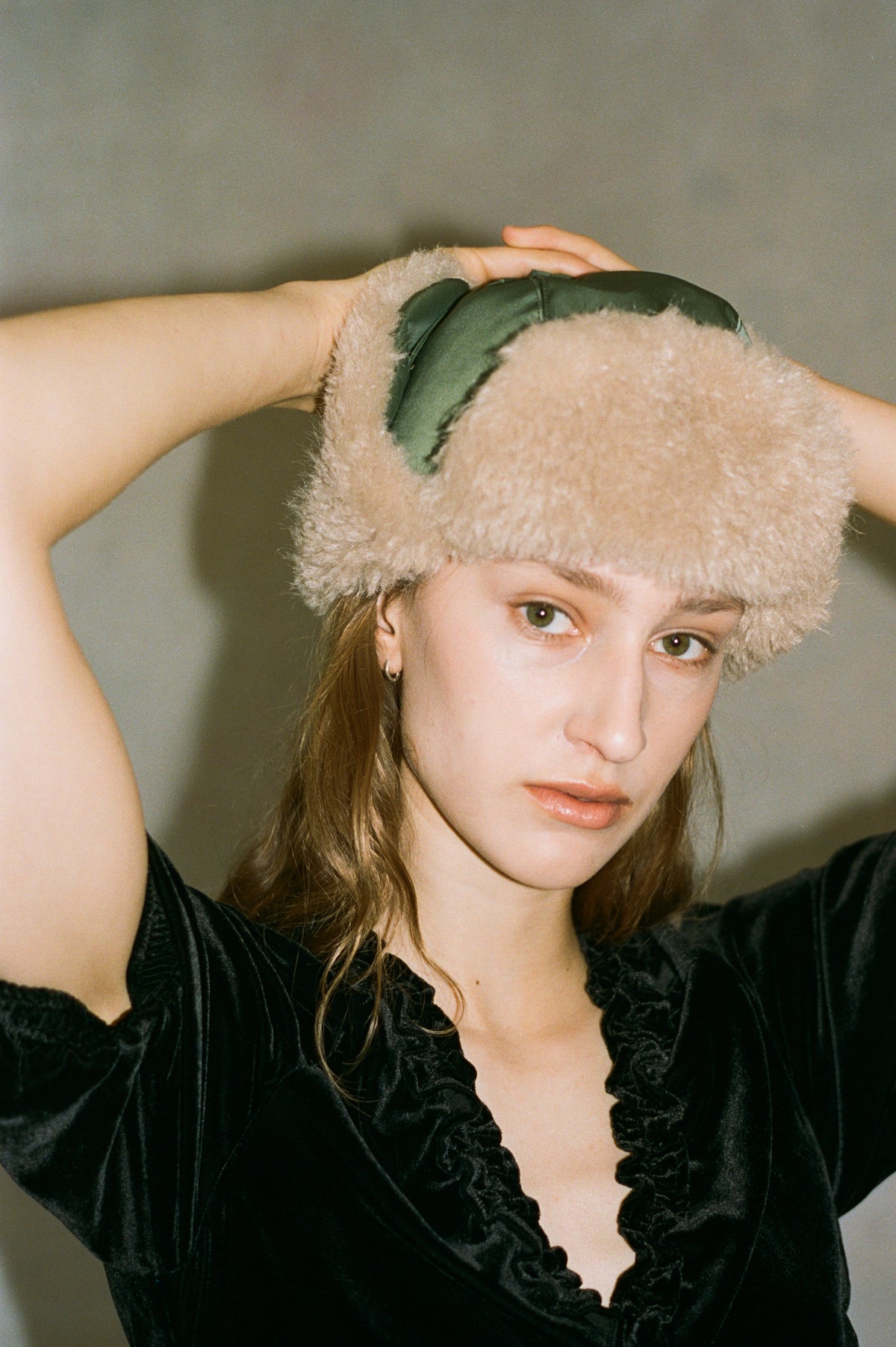 Close-up campaign portrait of a model adjusting a beige fuzzy hat with dark green panels, wearing a black velvet top with a ruffled neckline.