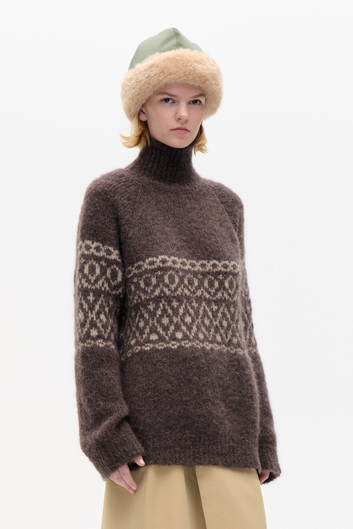 Model wearing a brown turtleneck sweater with cream geometric bands, beige trousers and a sage green hat with a fluffy brim.