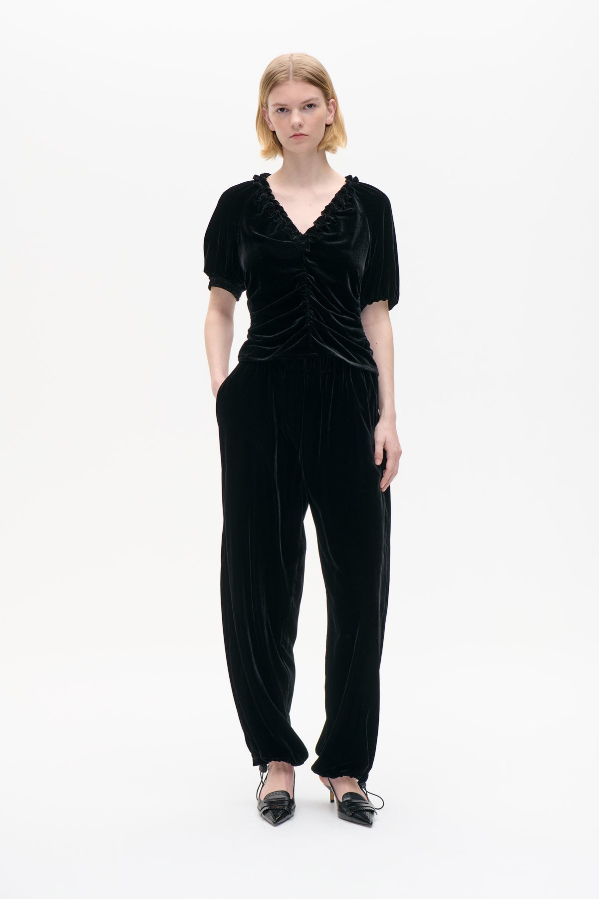 Front studio view of a model wearing a black velvet top with a ruffled V-neck, puff sleeves and ruched waist, with loose ankle-tie trousers.
