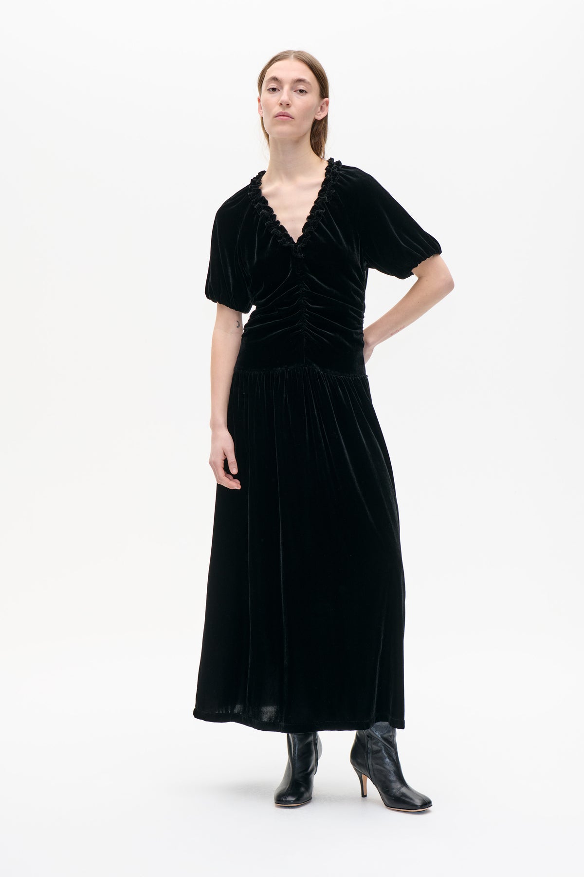 Model wearing a black velvet midi dress with puff sleeves, a ruffled V-neck and ruched waist, styled with black heeled boots.