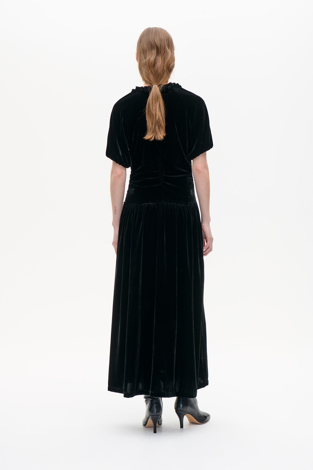 Back view of a black velvet maxi dress with puff sleeves, a ruffled neckline, gathered waist and flowing skirt.