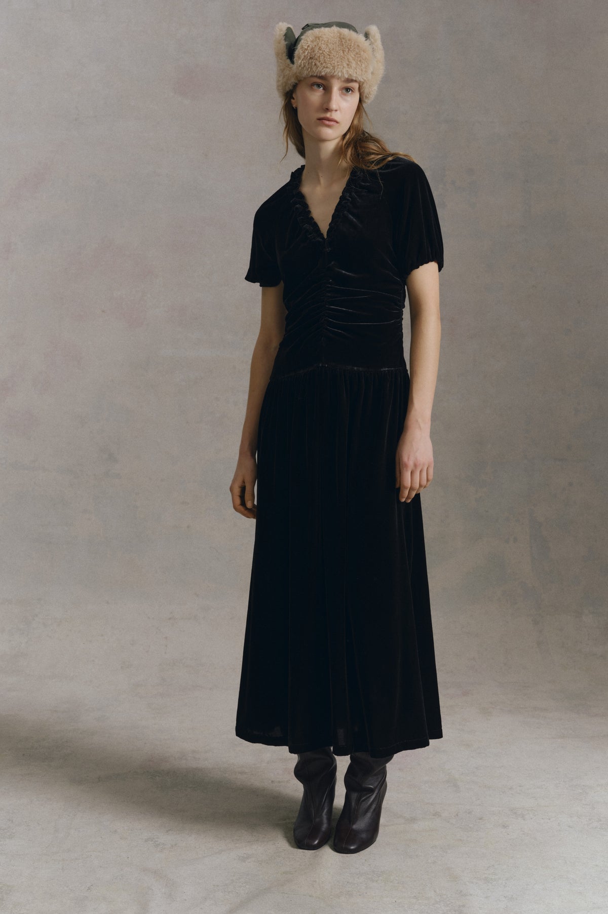 A model wears a long black dress with puff sleeves and a gathered neckline, paired with a cream fuzzy hat and black boots.