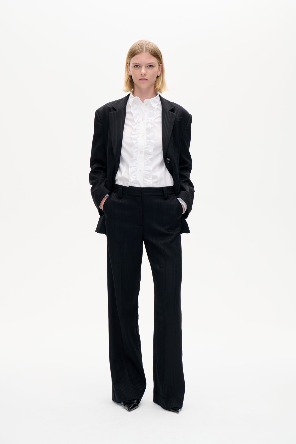 Mannequin de face portant un blazer noir à fines rayures et un pantalon large, associé à une chemise blanche boutonnée à volants, sur fond blanc uni.
