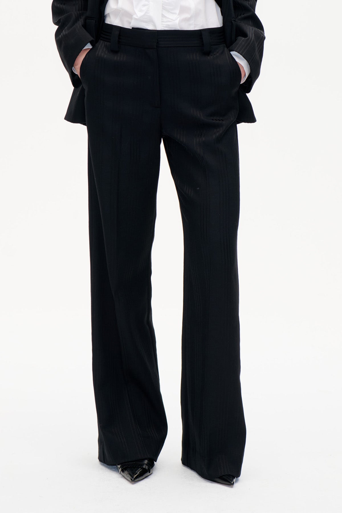Vue de face d'un pantalon noir à jambes larges, fines rayures verticales et taille haute, porté avec une chemise blanche et une veste noire.