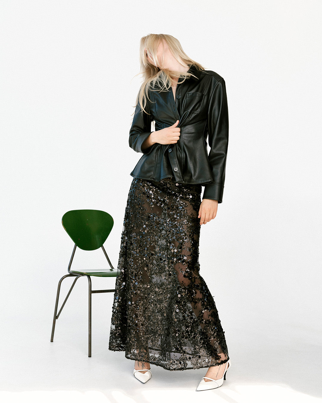 Mannequin en veste noire brillante boutonnée et jupe maxi noire transparente à sequins, debout près d'une chaise verte, chaussée d'escarpins blancs.