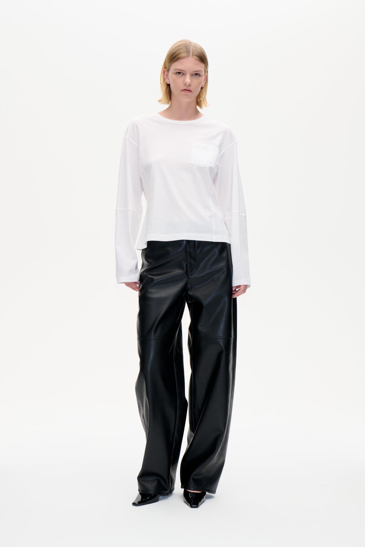 Model wearing a white long-sleeved crew-neck top with a chest pocket and black wide-leg trousers, shown from the front.