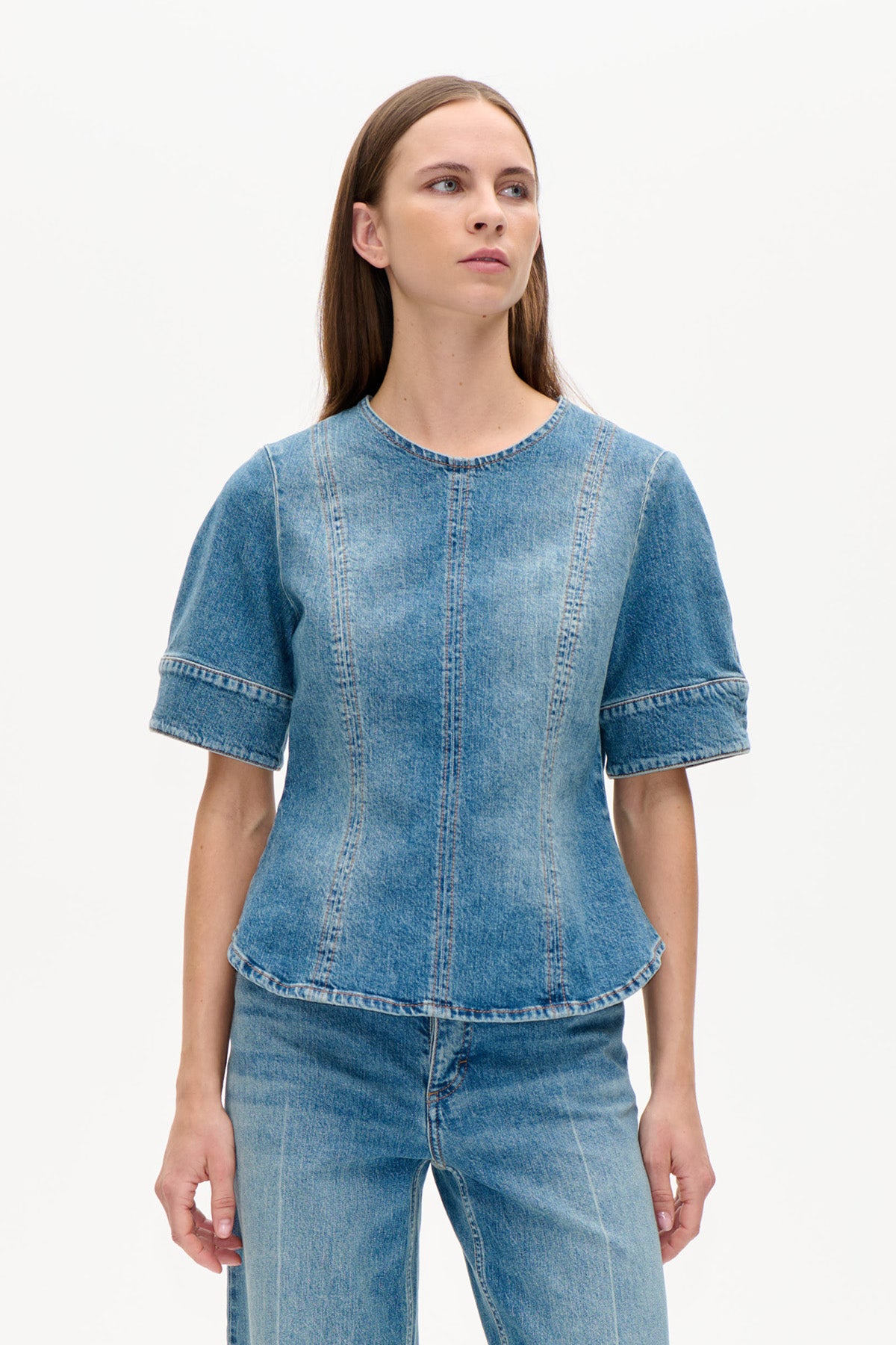 Vue de face d'un haut en denim bleu à encolure ronde, manches courtes et larges, coutures verticales formant des panneaux, porté avec un jean.