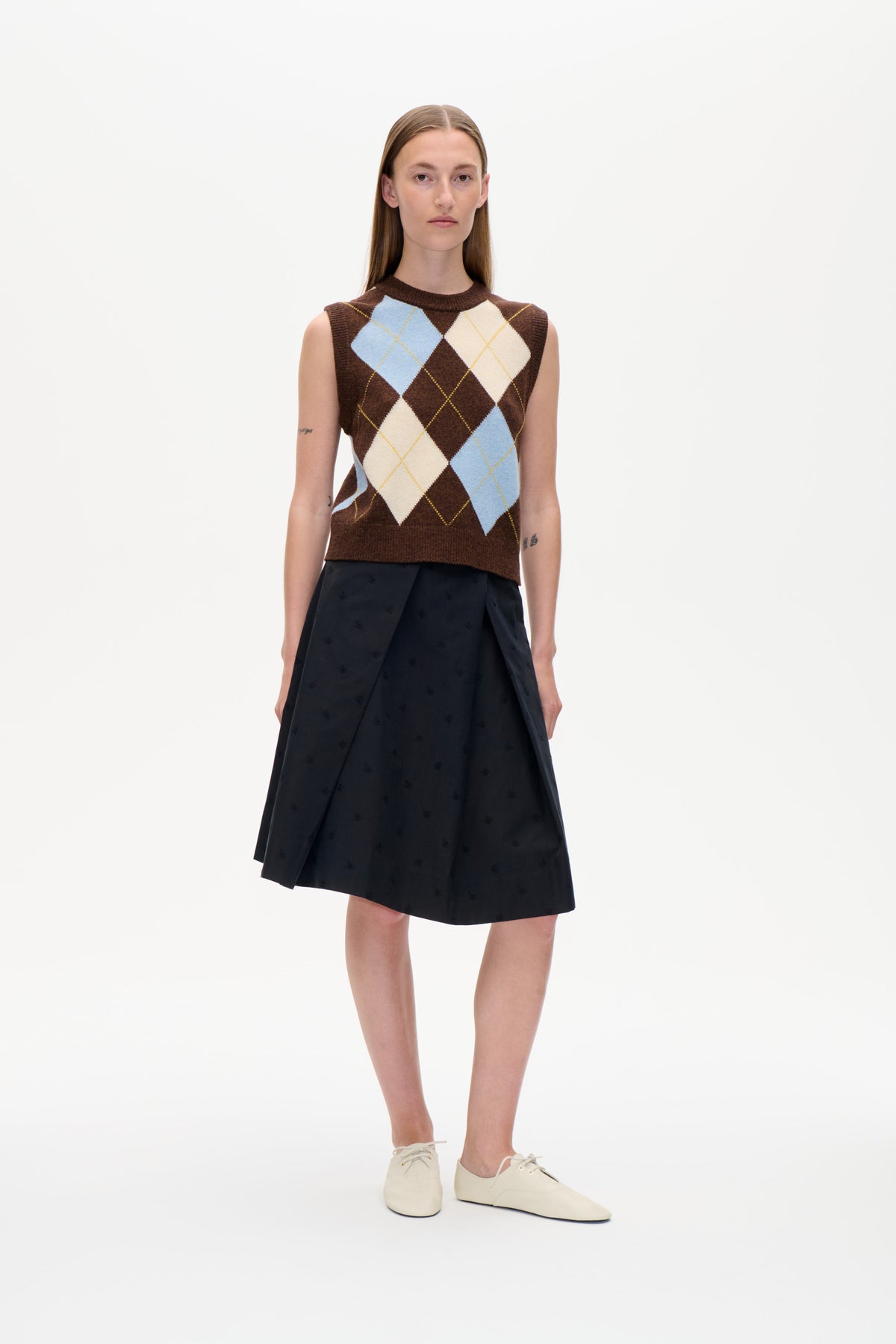 Front view of a model wearing a brown sleeveless knit vest with blue and cream argyle diamonds, paired with a black skirt.