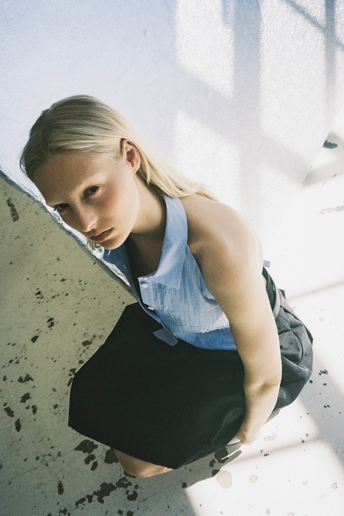 A model leans forward in a light blue sleeveless collared button-front top and black skirt, photographed from above.