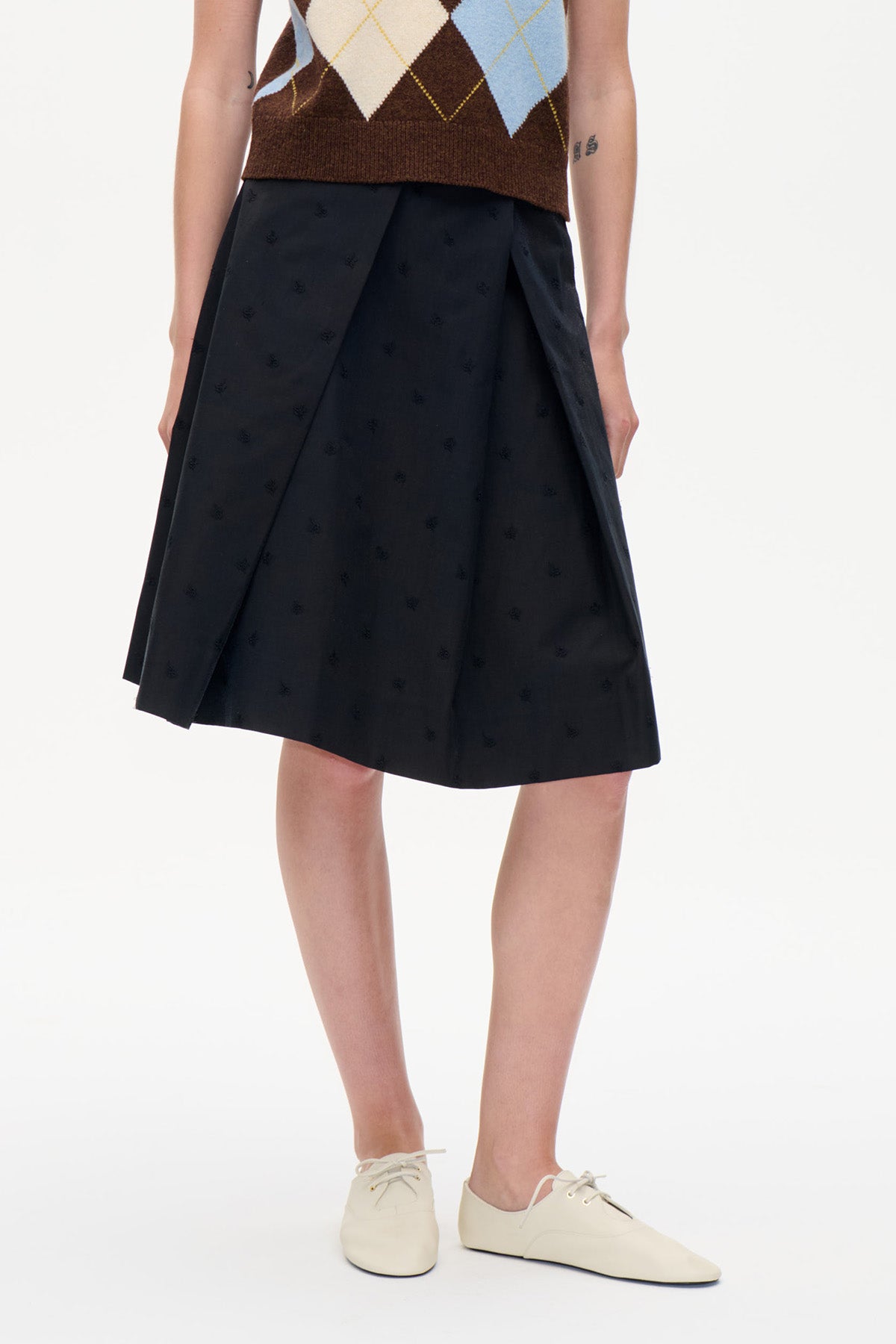 Close view of a black knee-length skirt with subtle tonal motifs and overlapping pleated front panels, styled with an argyle knit vest.