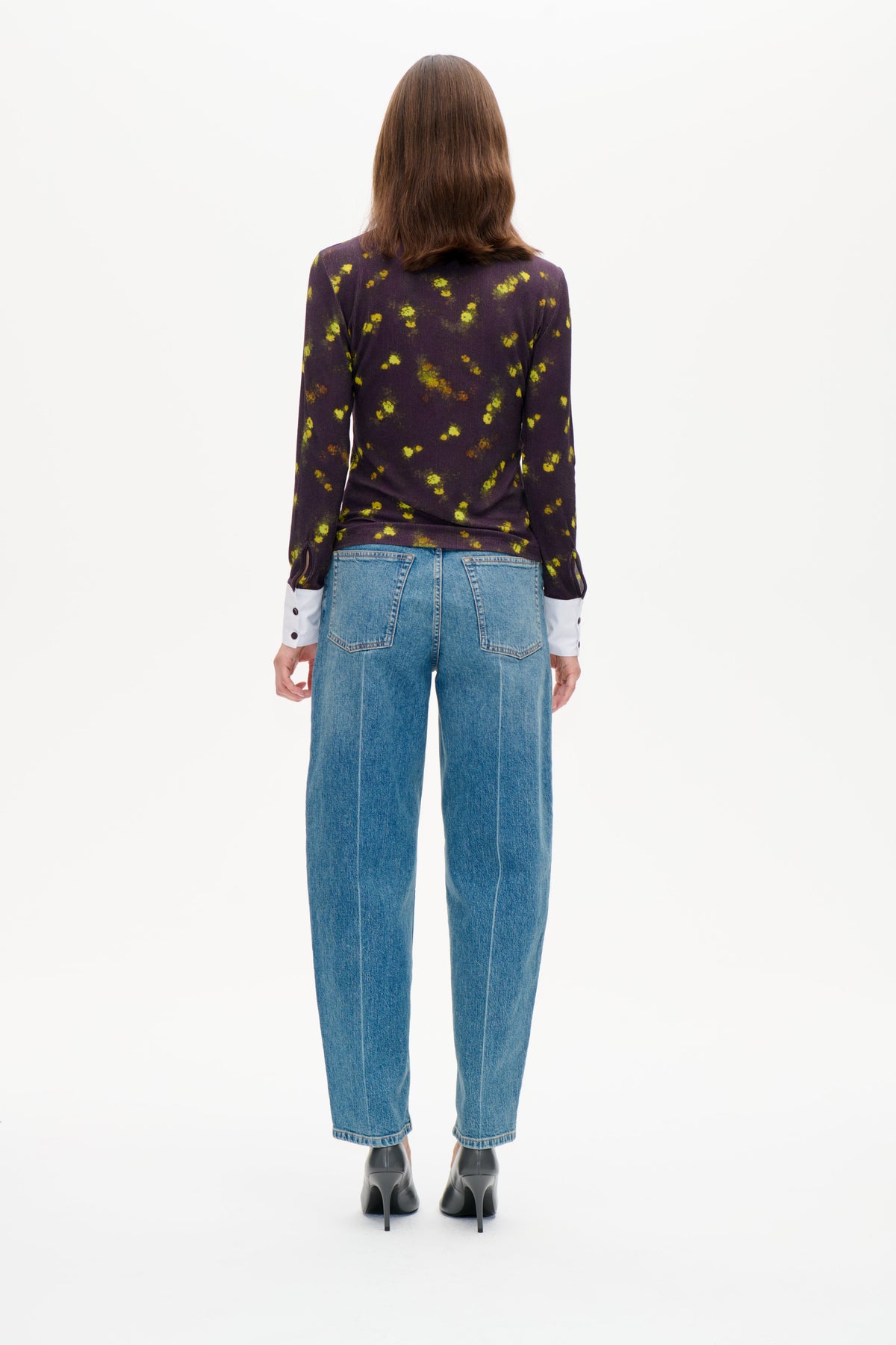 Back view of a model wearing a dark purple long-sleeved top with scattered yellow-green floral markings and blue jeans.