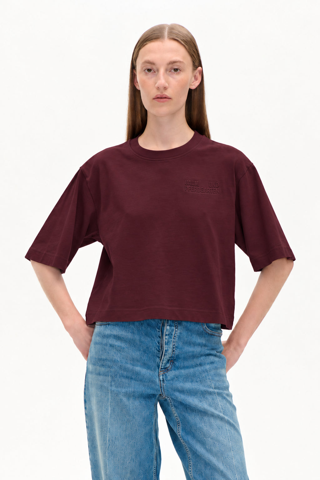 Model draagt een cropped bordeaux T‑shirt met korte mouwen, een borstlogo in reliëf en lichtblauwe jeans met patroon.