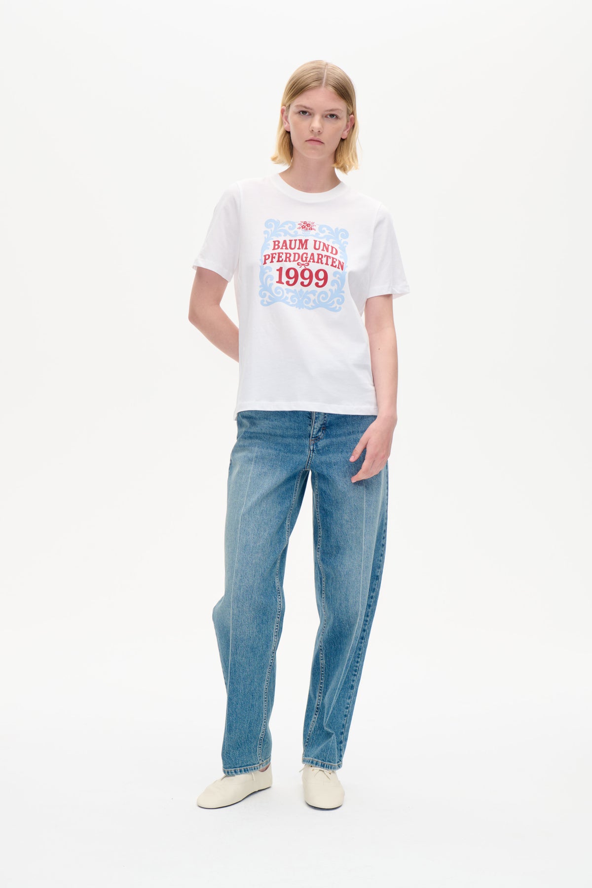 Model wearing a white short-sleeved T-shirt with a blue-and-red ornamental graphic reading “Baum und Pferdgarten 1999,” styled with blue jeans.