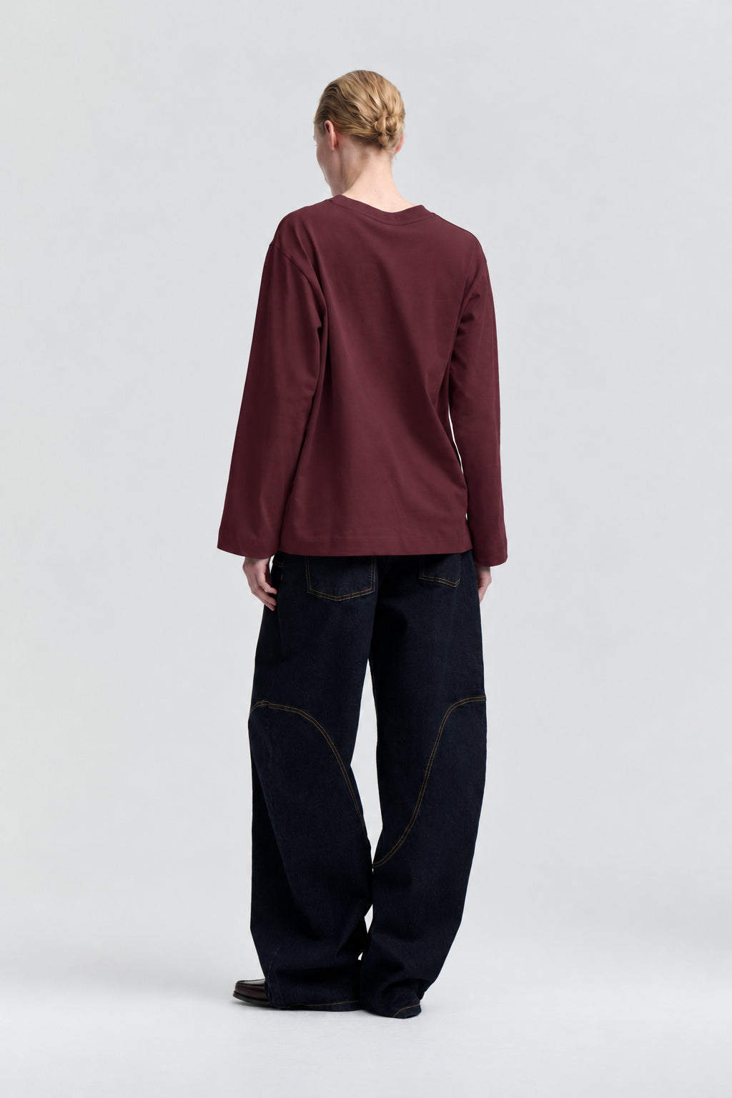 Achteraanzicht van een model dat een effen bordeauxrode top met ronde hals en lange mouwen draagt, met donkere wijde jeans.