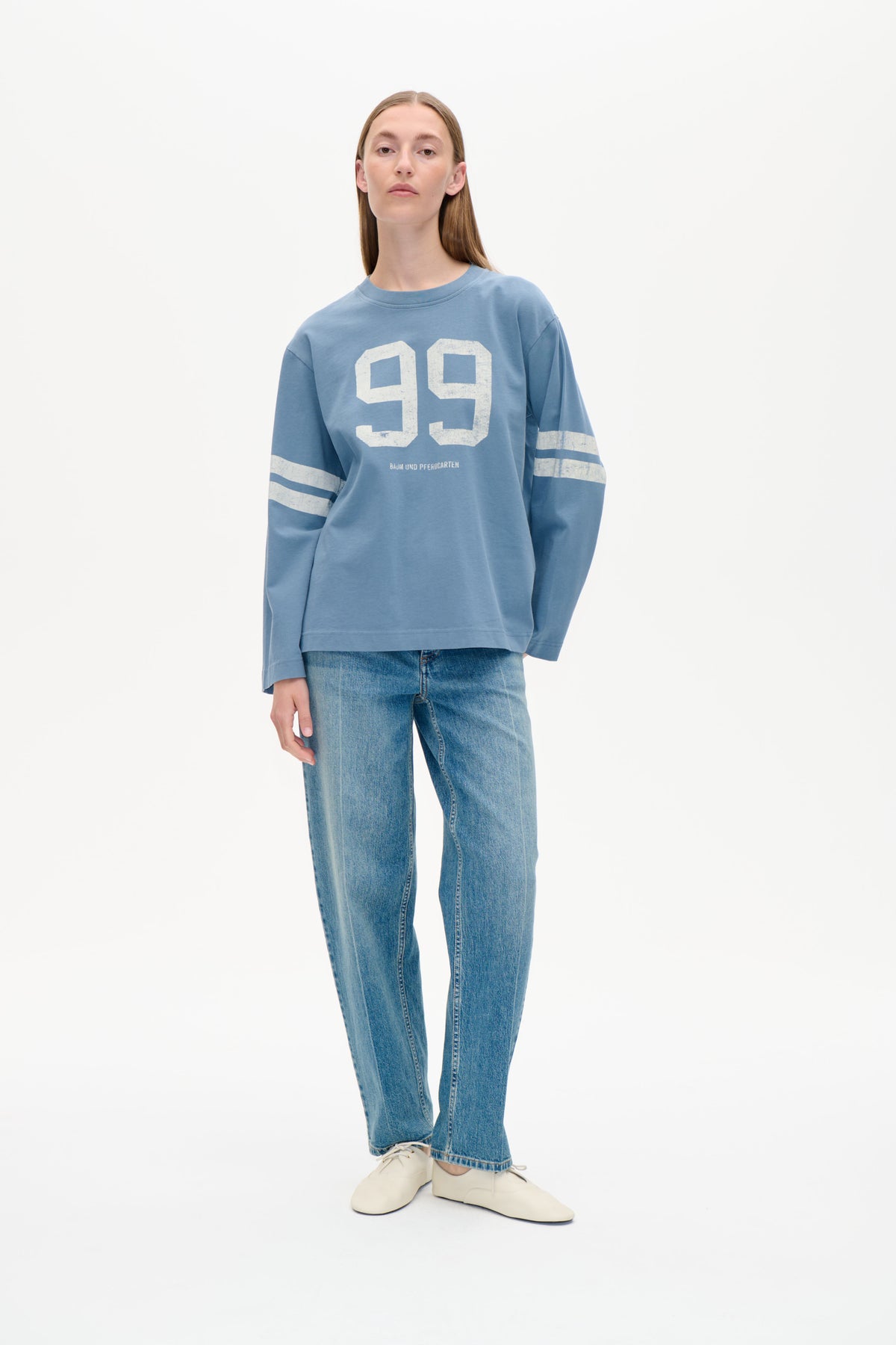 Vorderansicht eines Models, das ein lockeres blaues Sweatshirt mit großem, leicht verwittertem weißen 99-Print und gestreiften Ärmeln zu Jeans trägt.