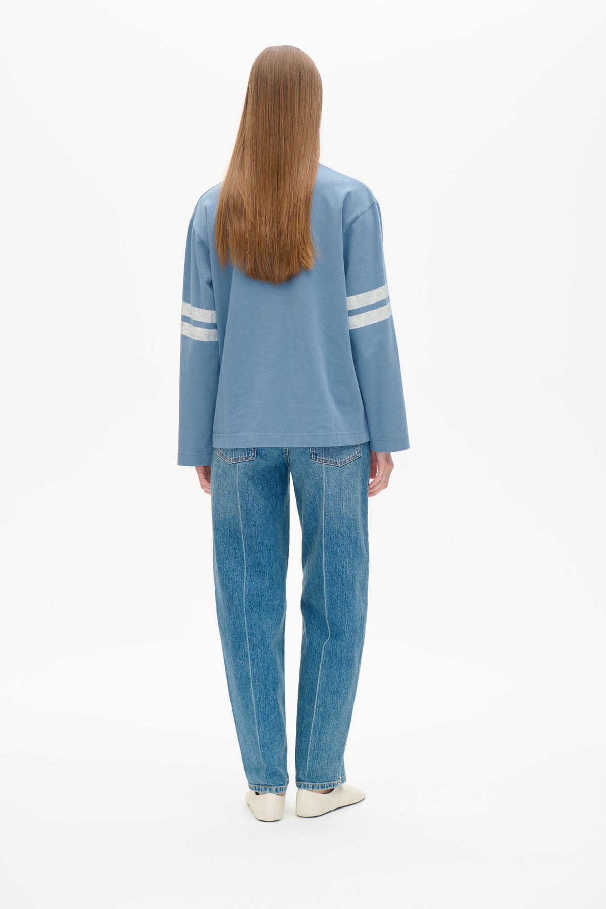Rückenansicht eines Models, das ein hellblaues Langarm-Sweatshirt mit weißen Streifen an den Ärmeln und eine blaue Jeans trägt.