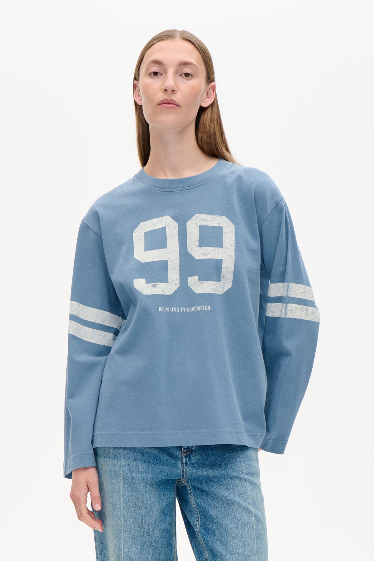 Model trägt ein staubblaues Sweatshirt mit langen Ärmeln, weißem '99'-Print auf der Brust und zwei Streifen an den Ärmeln, kombiniert mit Jeans.