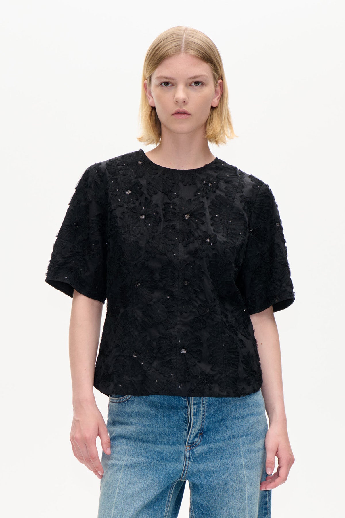 Front view of a black short-sleeved top with raised floral detailing and small embellishments, paired with blue jeans.