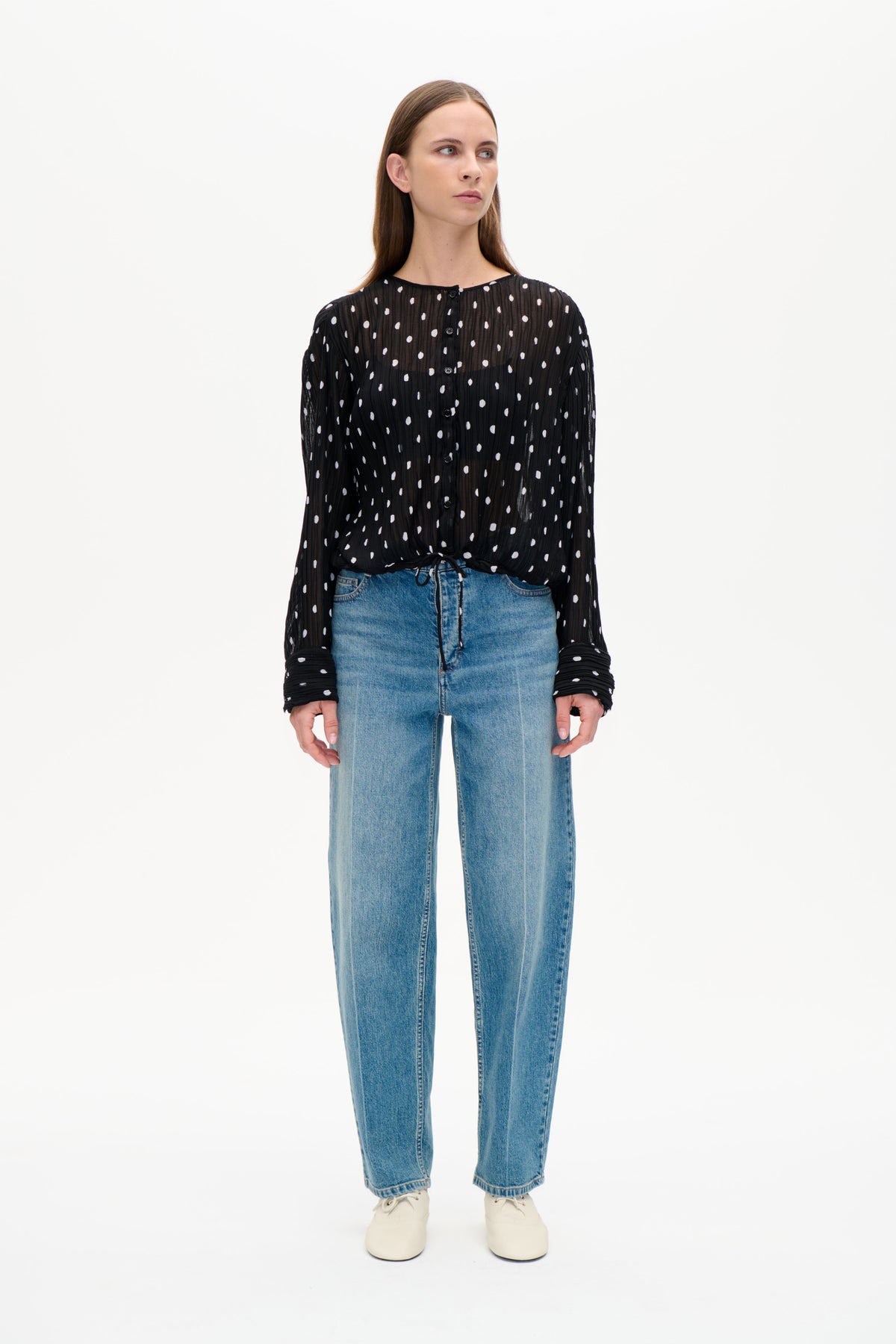 Vorderansicht einer schwarzen, transparenten Bluse in Knitter-Optik mit verstreuten weißen Punkten, durchgehender Knopfleiste, langen Ärmeln und Kordelzug am Saum.