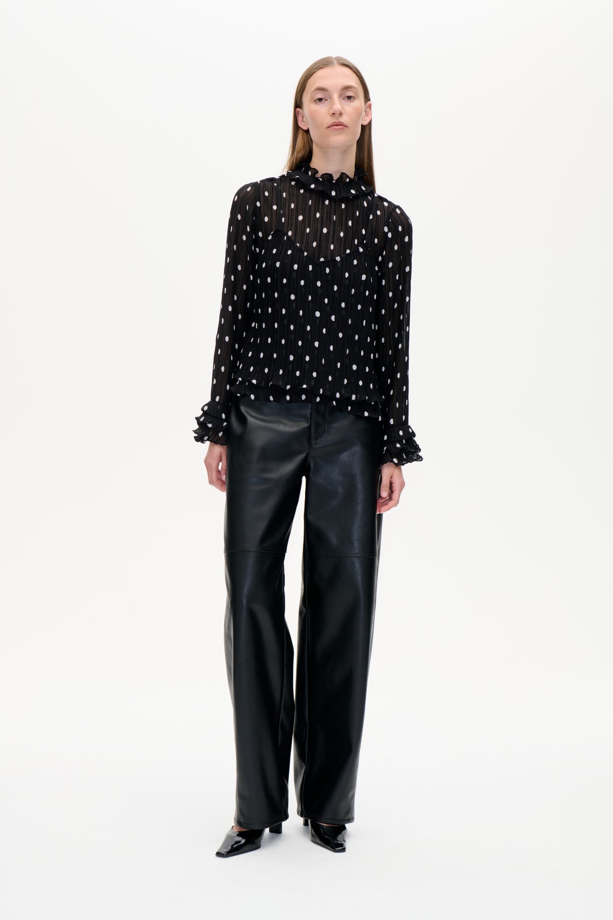 Model van voren draagt een transparante zwarte blouse met witte stippen en ruches, gecombineerd met een zwarte broek met wijde pijpen.