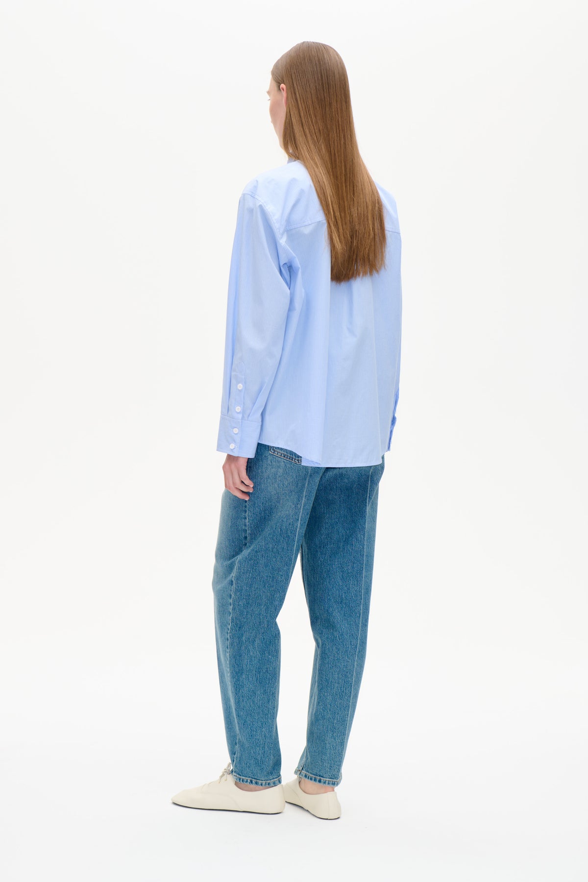 Back view of a model wearing a light blue fine-striped button-up shirt with long sleeves and blue jeans.
