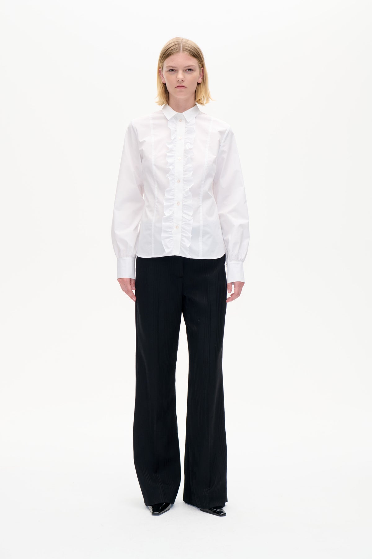 Front-facing studio portrait of a model wearing a white ruffle-front button-up shirt and black wide-leg trousers.