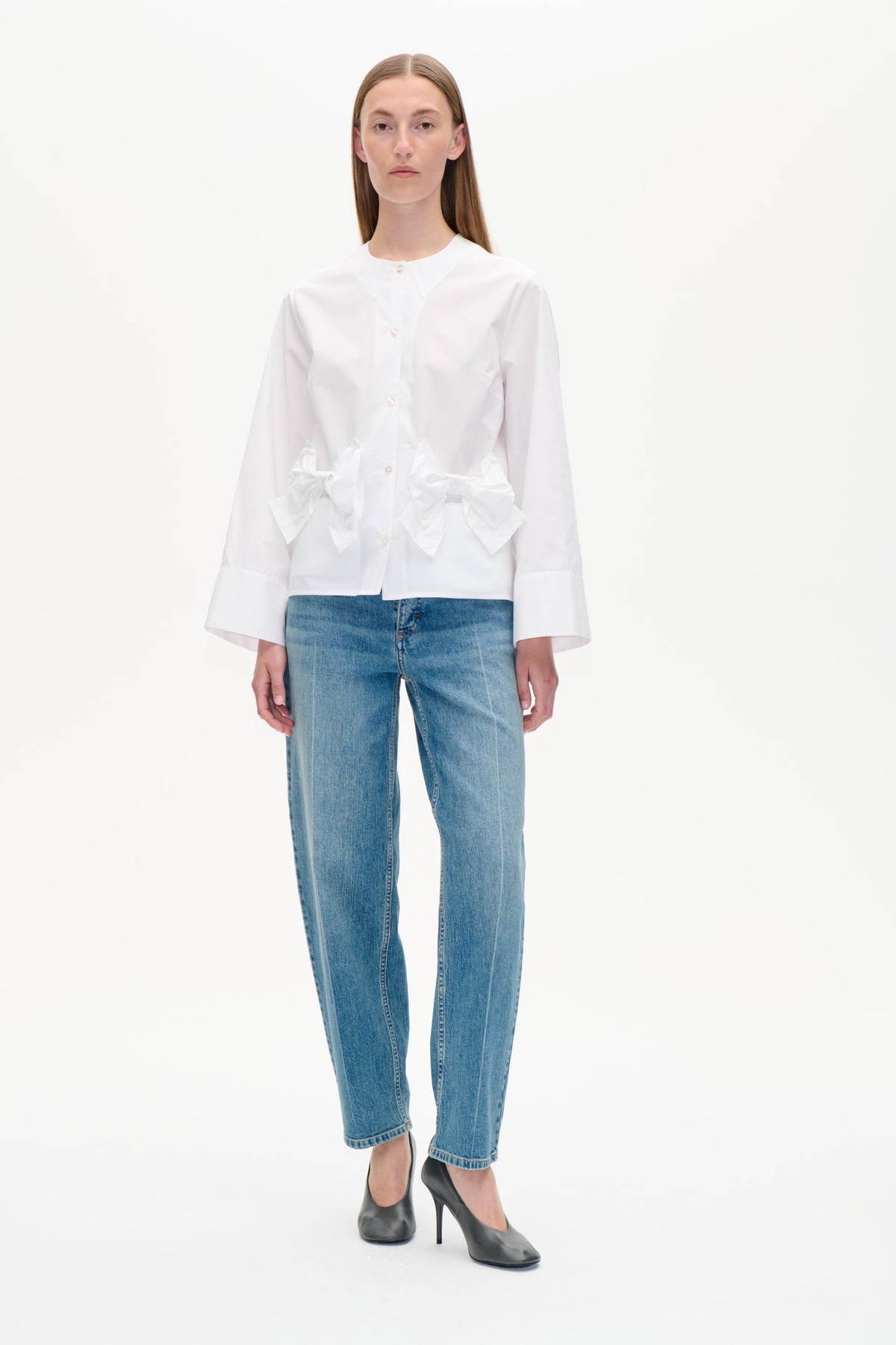 Vue de face d'une chemise blanche boutonnée à l'avant, avec un col pointu oversized et des nœuds sculpturaux à la taille, portée avec un jean bleu.