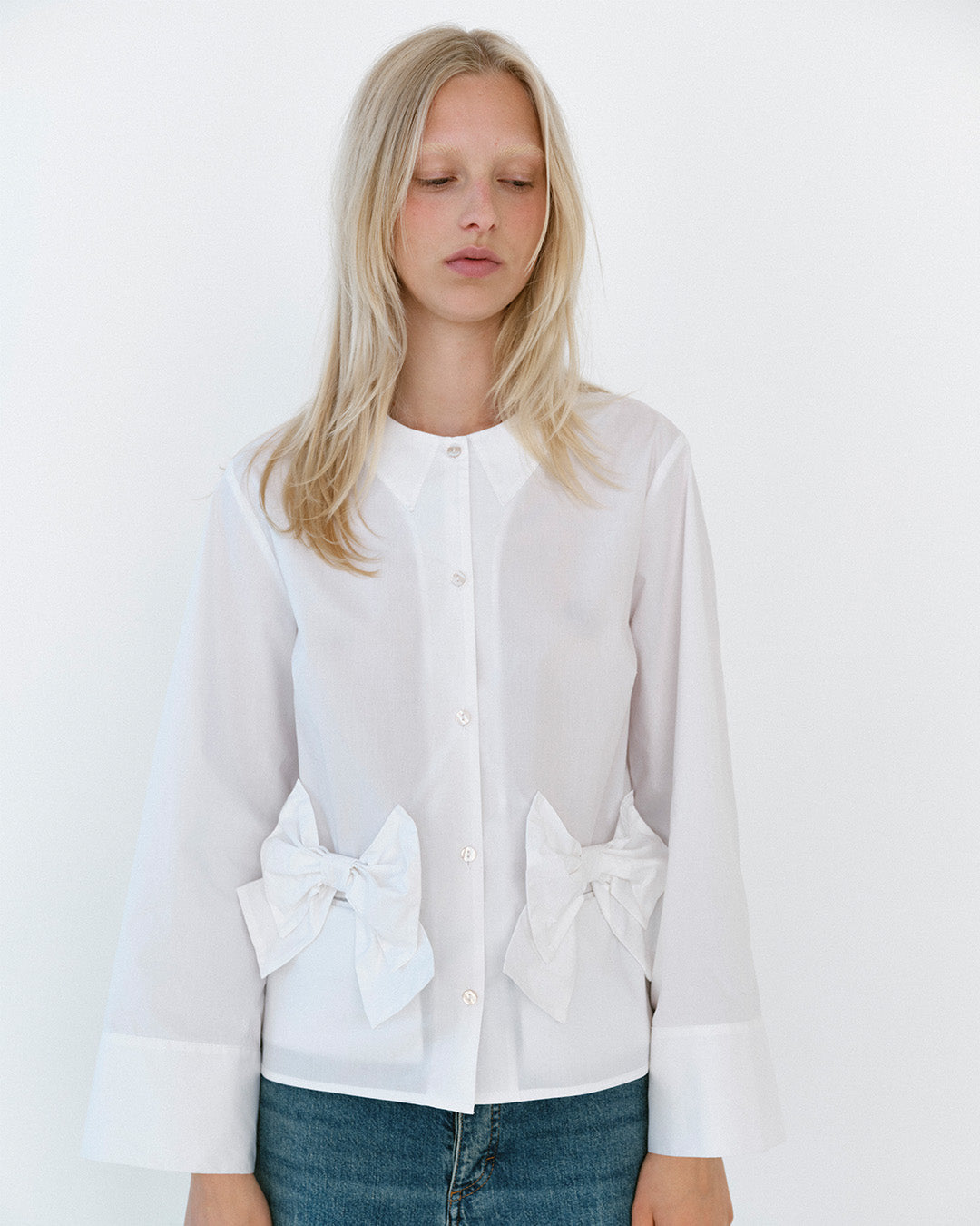 Mannequin portant une blouse blanche boutonnée à l'avant, col pointu, détails de nœuds oversized à la taille et poignets larges, portée avec un jean bleu.