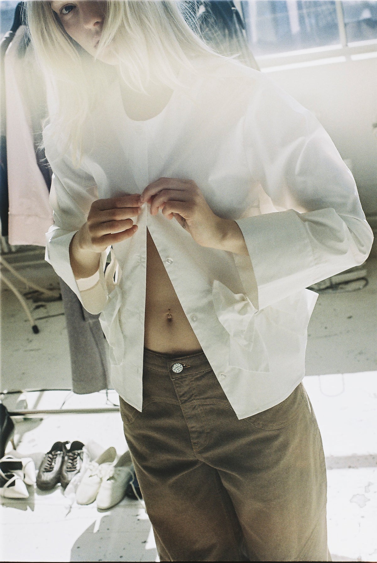 Portrait de campagne recadré d'un mannequin ajustant une chemise blanche oversized boutonnée sur un pantalon marron.