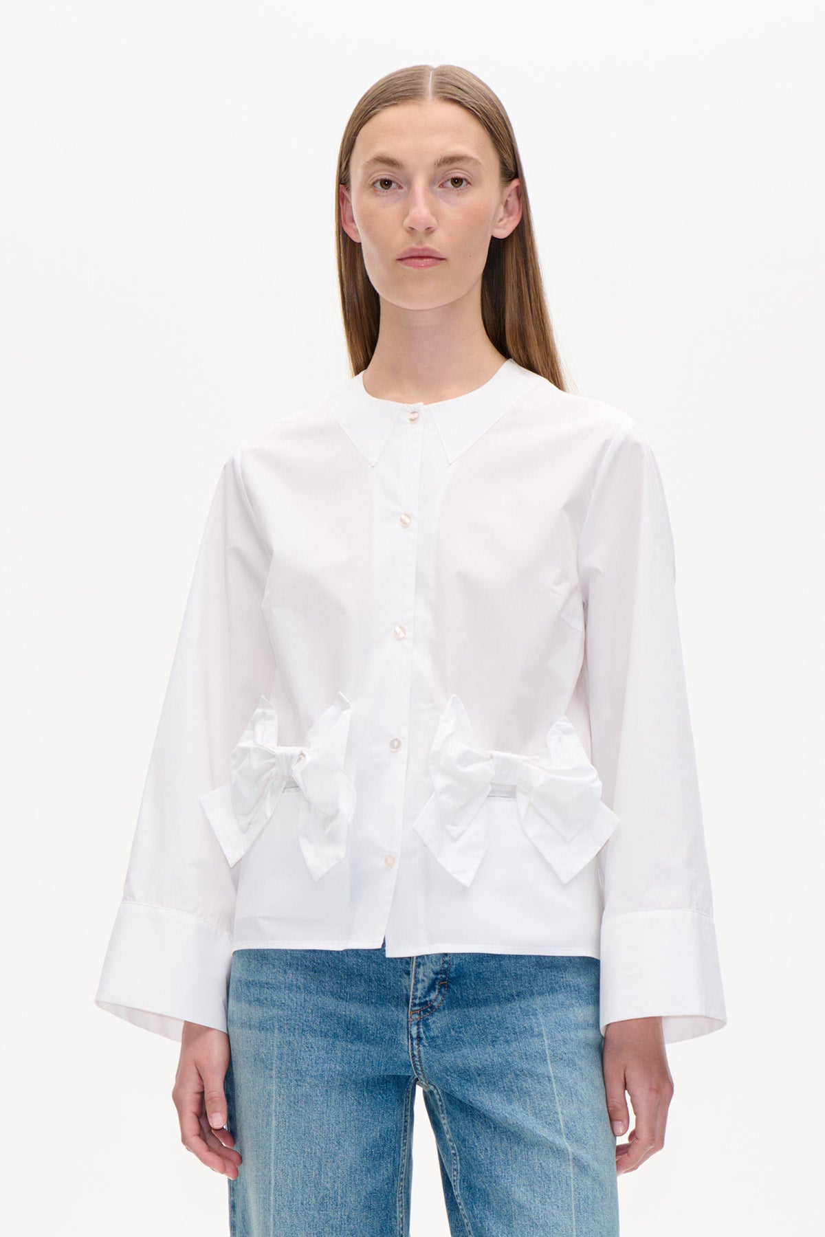 Vue de face d'une chemise blanche boutonnée à col pointu, poignets oversized et grands nœuds à la taille, portée avec un jean bleu.