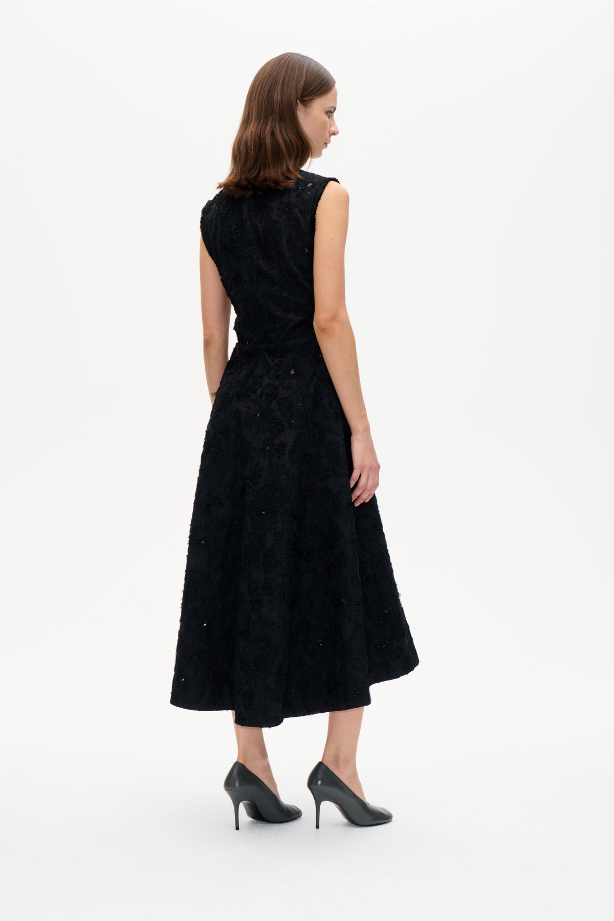 Model wearing a black sleeveless flared dress with raised floral detailing and scattered embellishments, shown from a side-back angle.