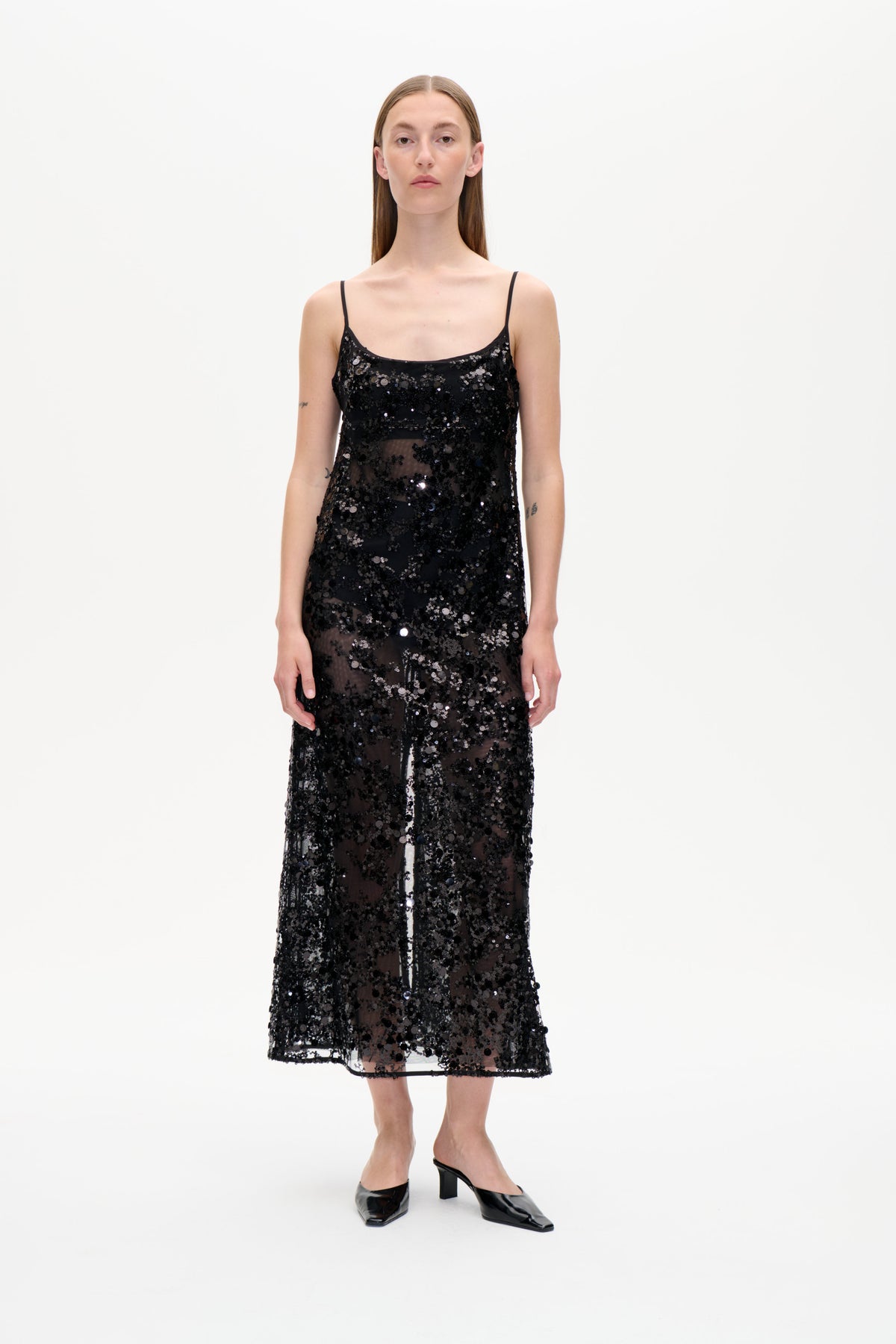 Front view of a black sleeveless slip dress with thin straps and dense sequins over sheer mesh, modeled against a white background.