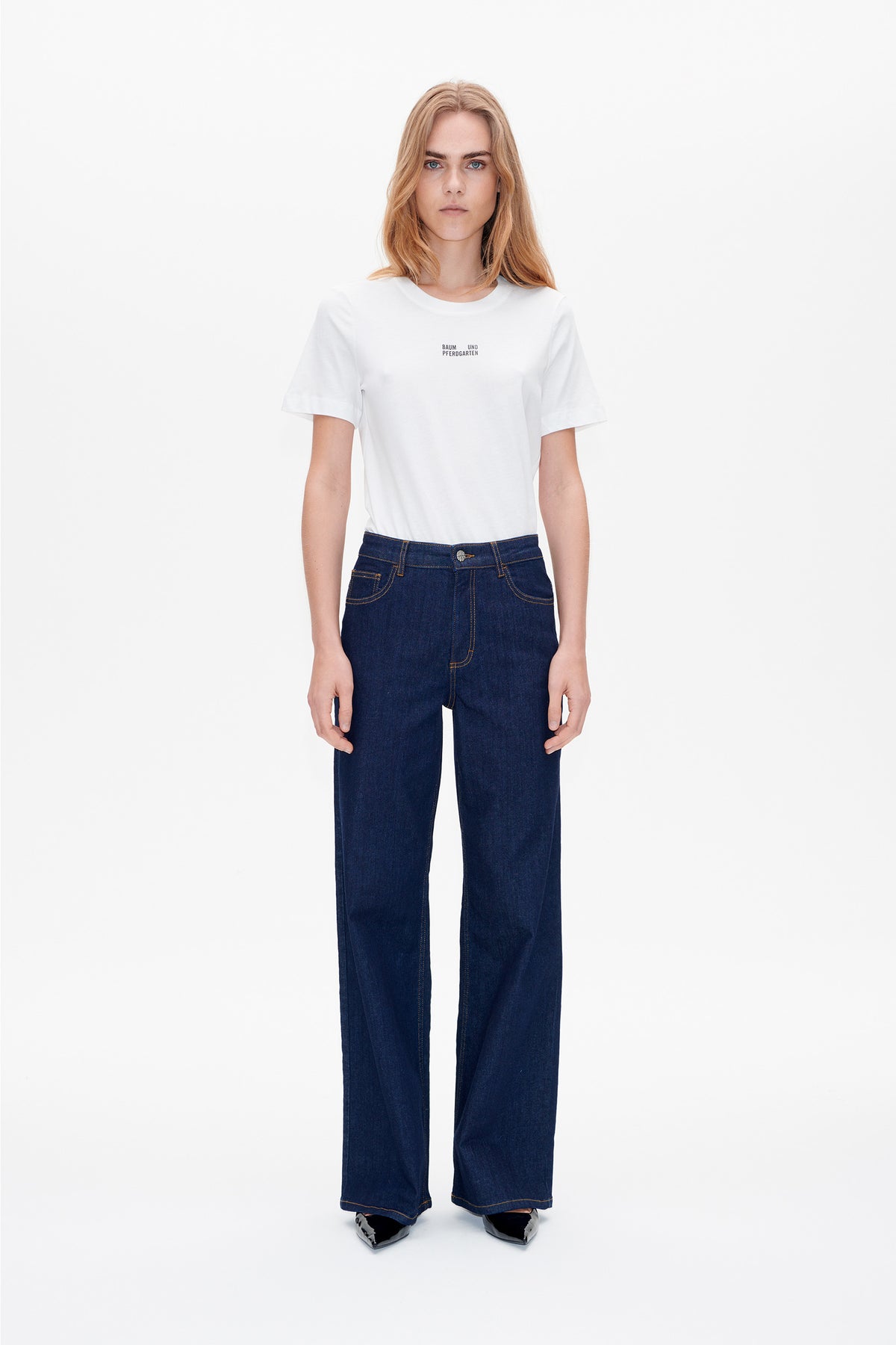 Front view of a model wearing a white crew-neck T-shirt with small Baum und Pferdgarten chest lettering and dark indigo wide-leg jeans.
