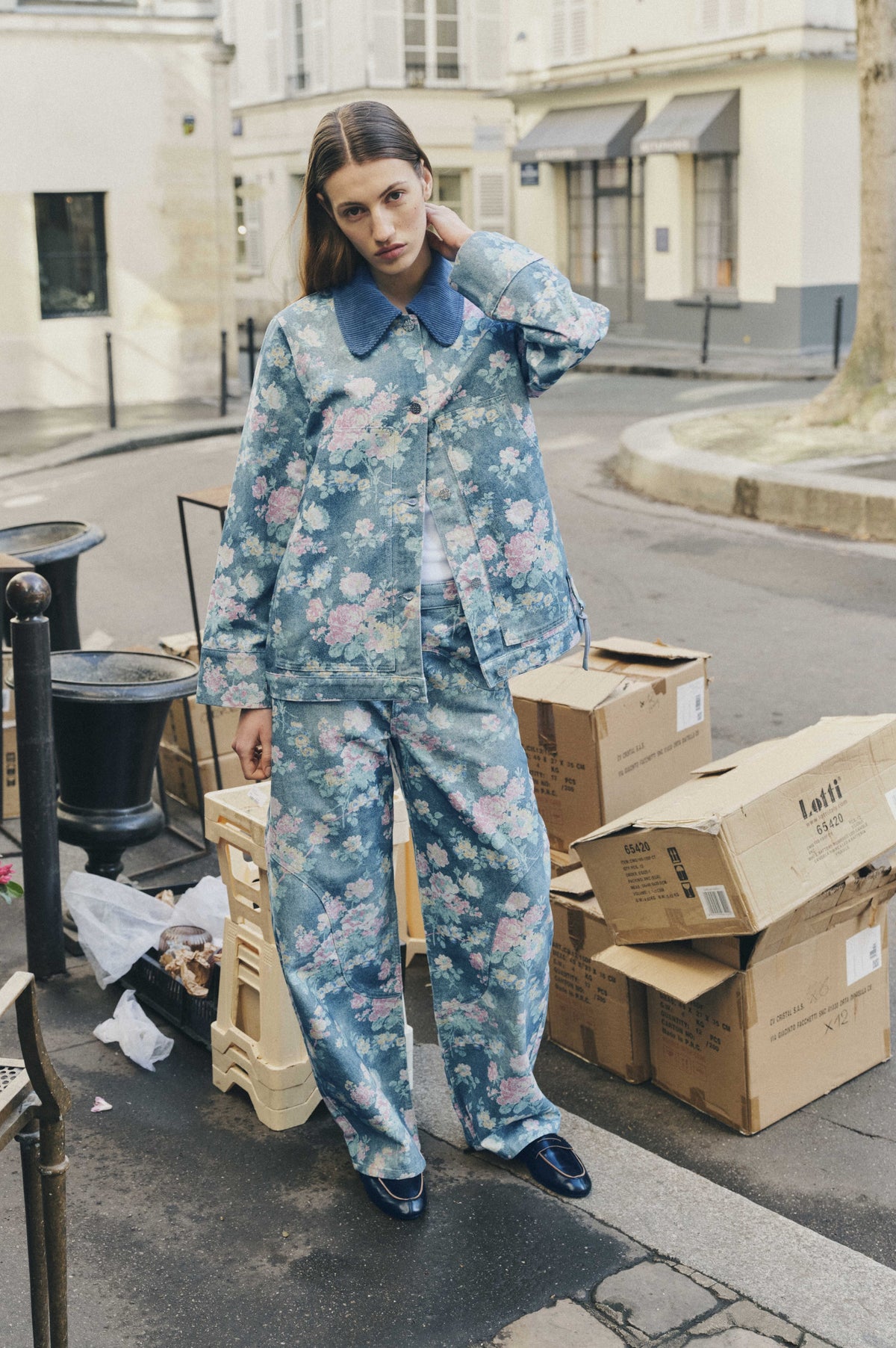 A model poses among stacked crates wearing a blue floral-print jacket with a wide ribbed collar and matching wide-leg trousers.
