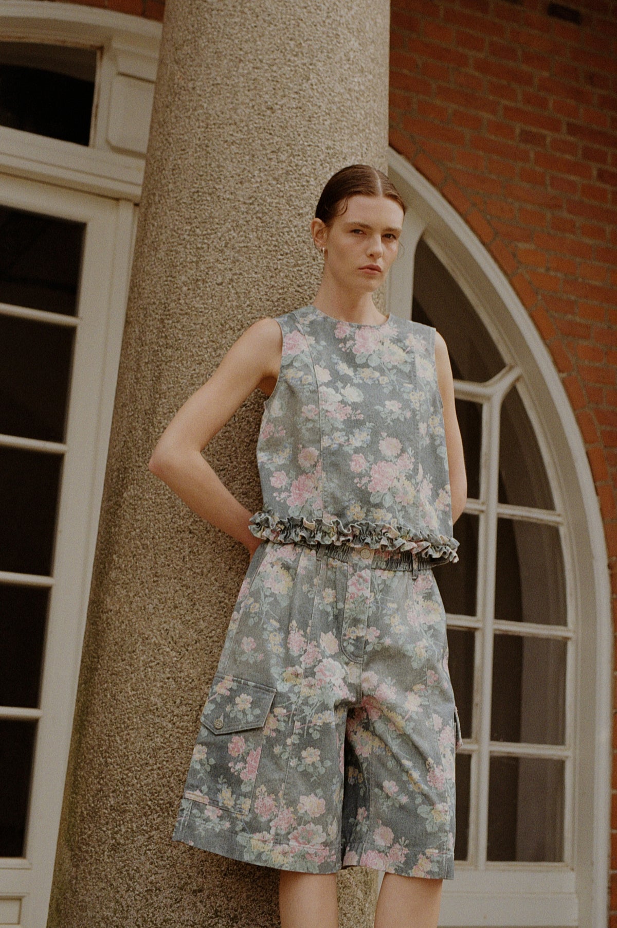 Model wearing a grey-blue floral sleeveless top with a ruffled waist and matching shorts, posed before brick and stone arches.