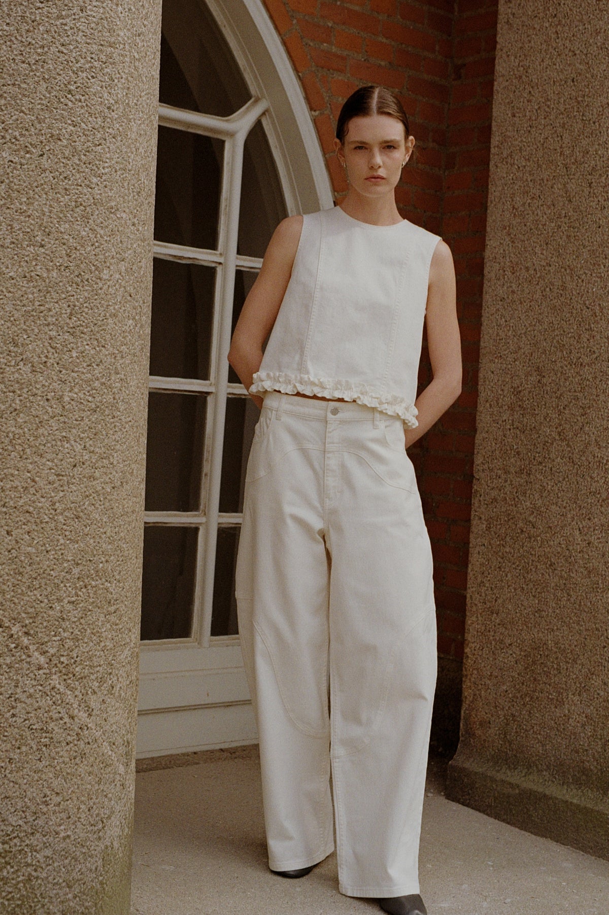 A model stands by a brick wall and window wearing a white sleeveless top with a ruffled hem and wide-leg white jeans.